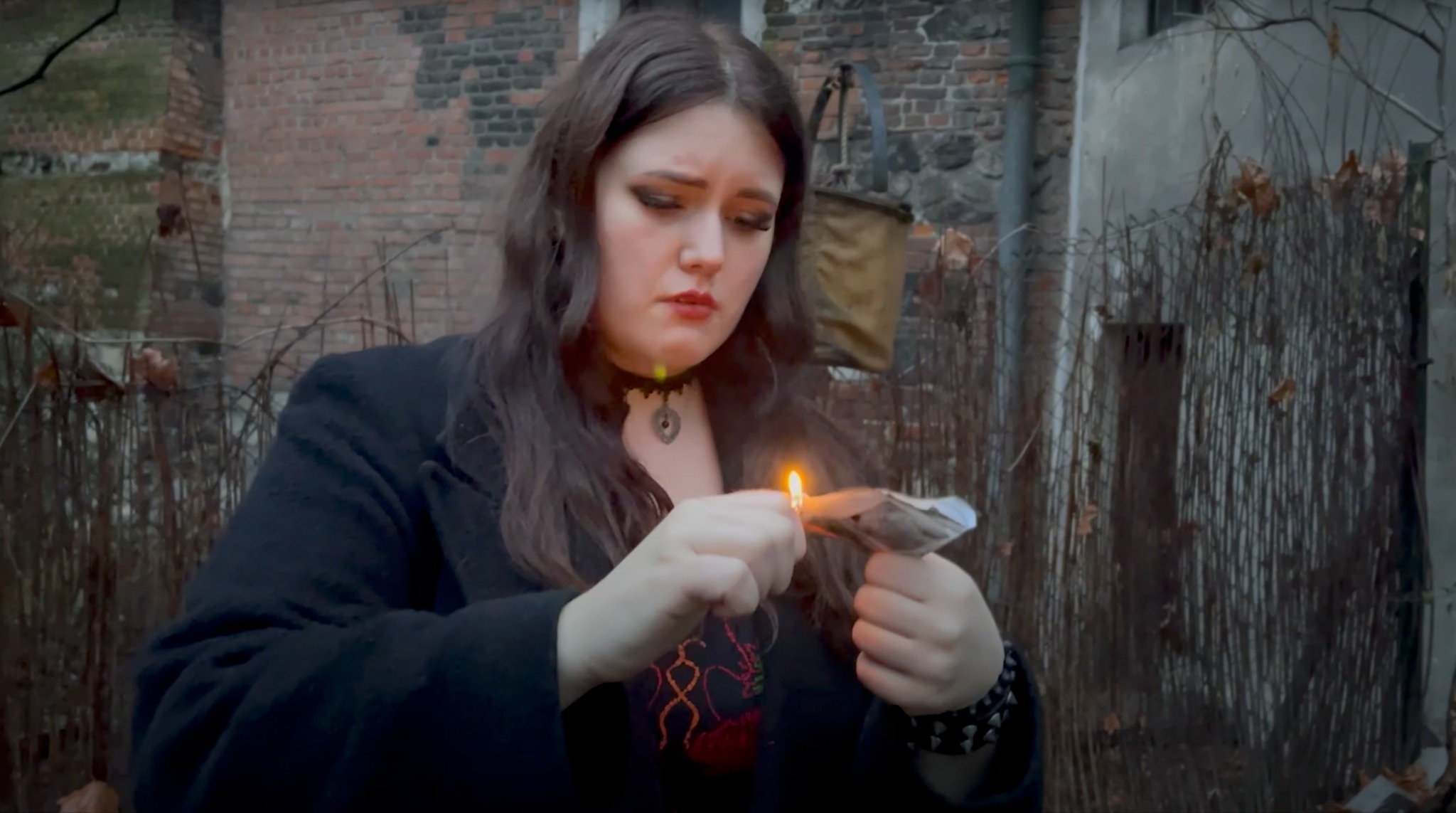 Młoda kobieta w czarnym płaszczu podpala banknoty zapalniczką na tle ceglanego budynku.