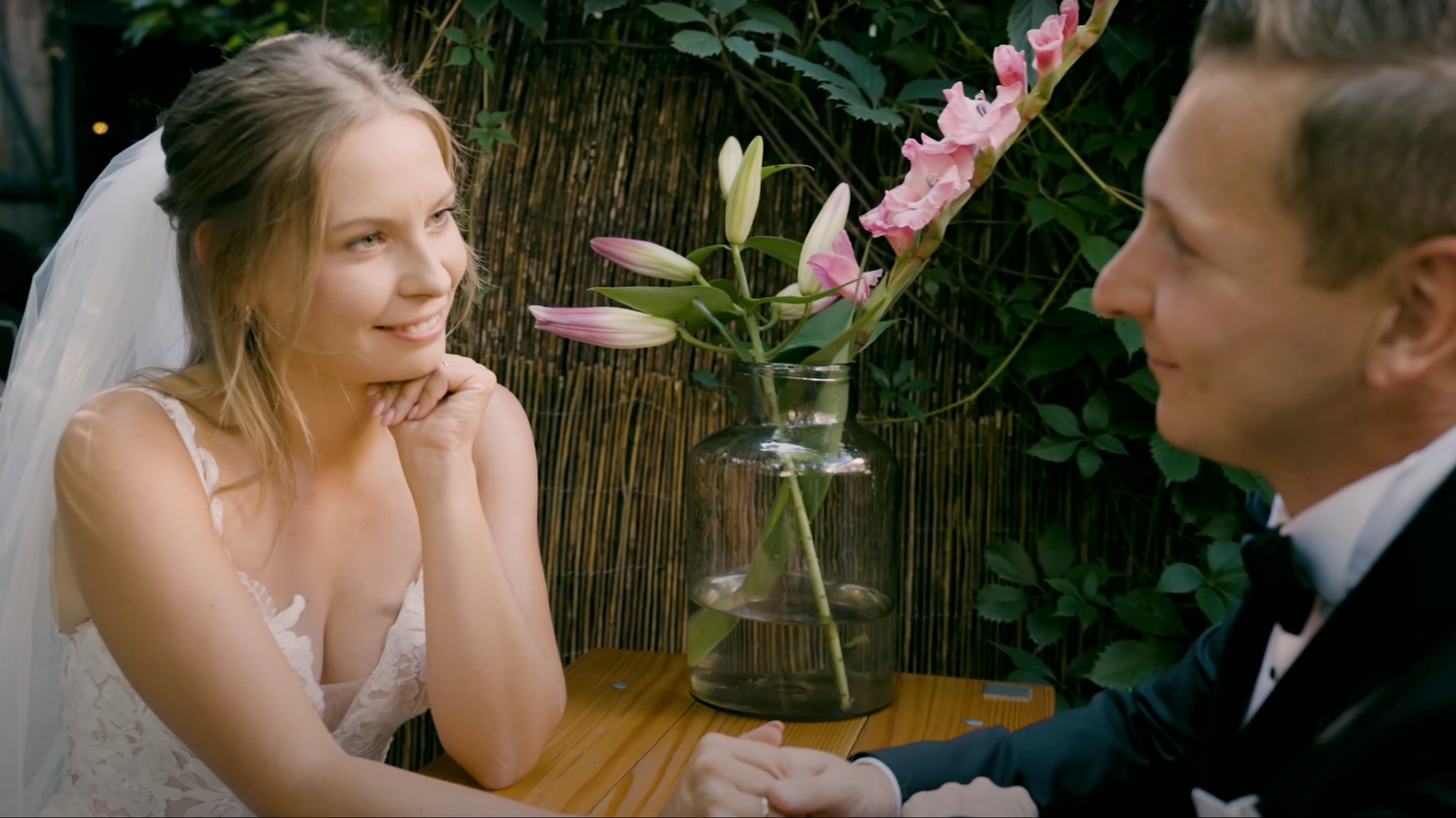 Panna młoda w koronkowej sukni spogląda z uśmiechem na pana młodego podczas kameralnego przyjęcia weselnego, z bukietem lilii i mieczyków w szklanym wazonie na drewnianym stole.