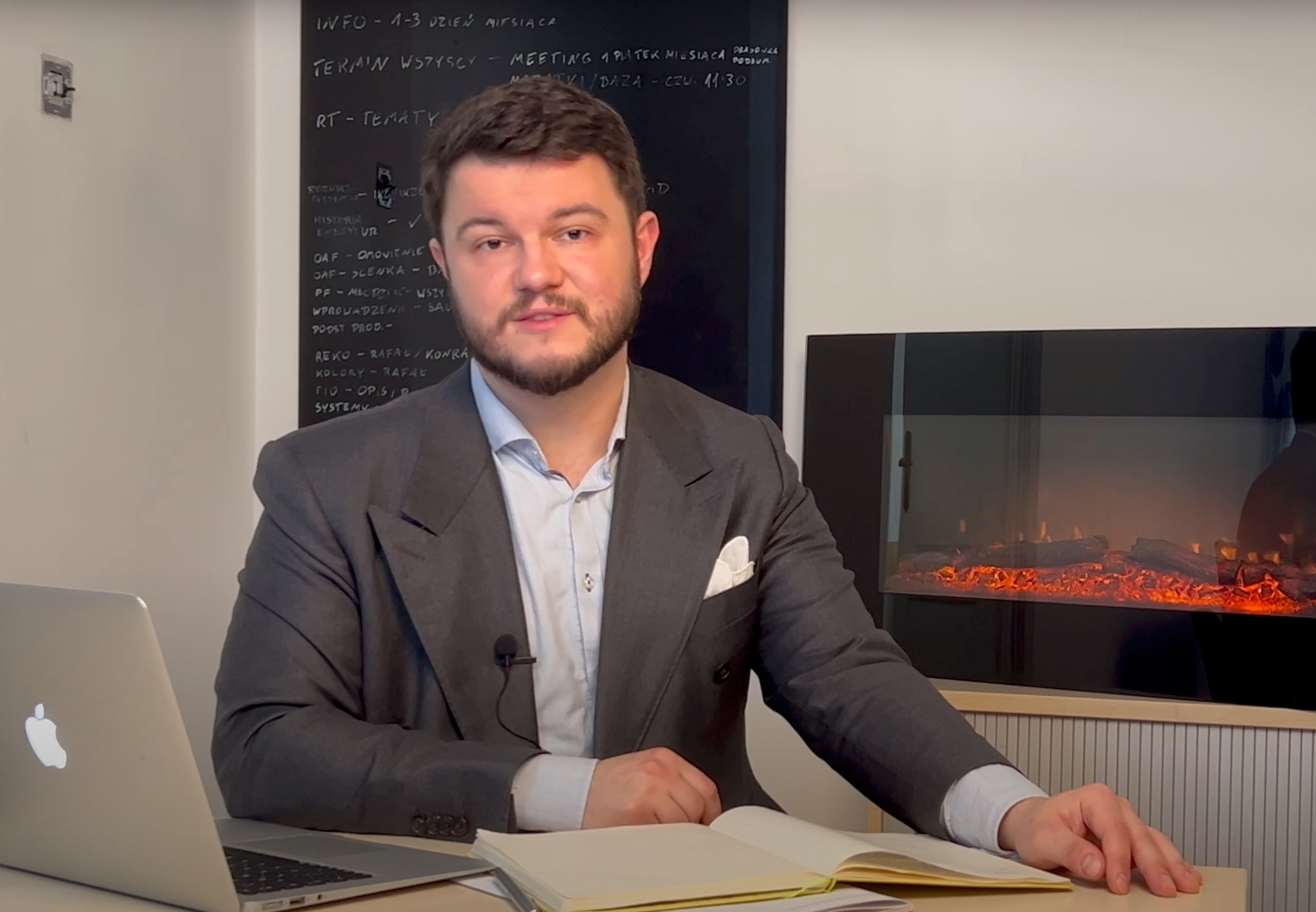 Mężczyzna w szarym garniturze siedzi przy biurku z laptopem i otwartym notatnikiem, w tle tablica z notatkami i imitacja kominka.