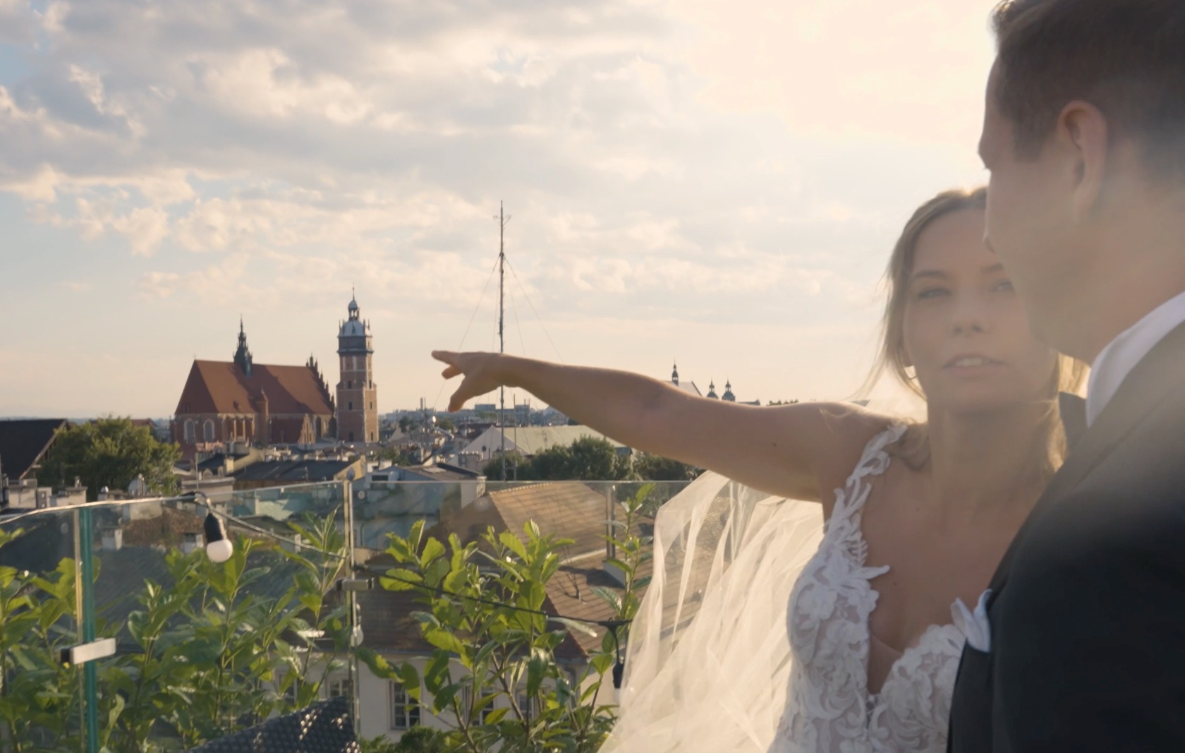 Panna młoda w koronkowej sukni i welonie wskazuje na panoramę miasta z kościołami podczas sesji ślubnej na dachu.