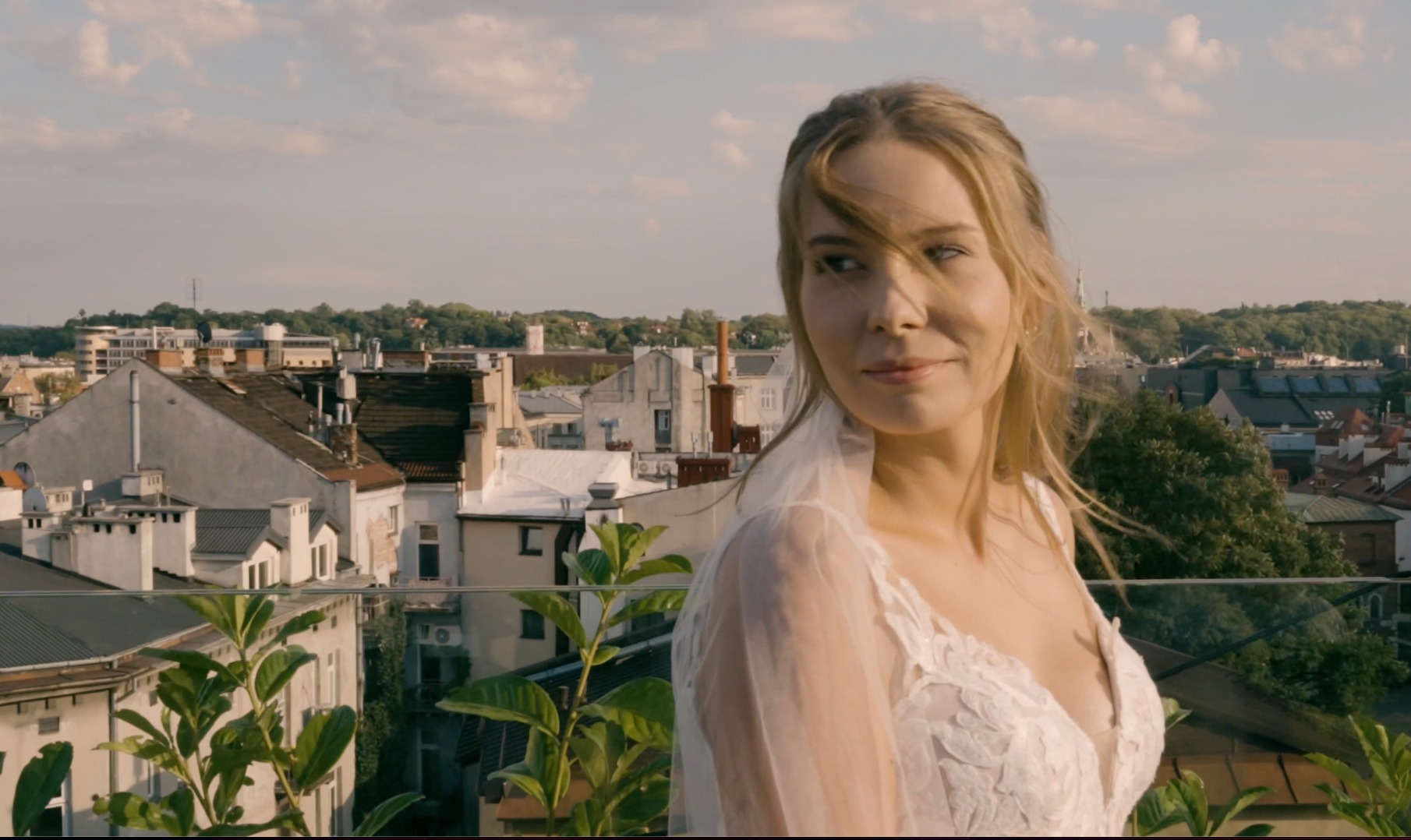 Panna młoda w koronkowej sukni i welonie na dachu budynku z widokiem na miasto w słoneczny dzień.