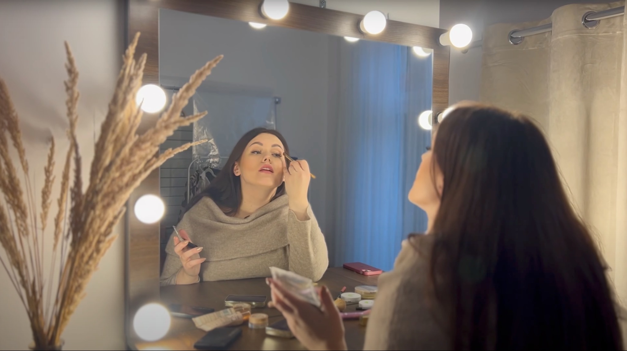Kobieta maluje rzęsy przed lustrem z żarówkami, na stole kosmetyki i dekoracyjna trawa pampasowa.