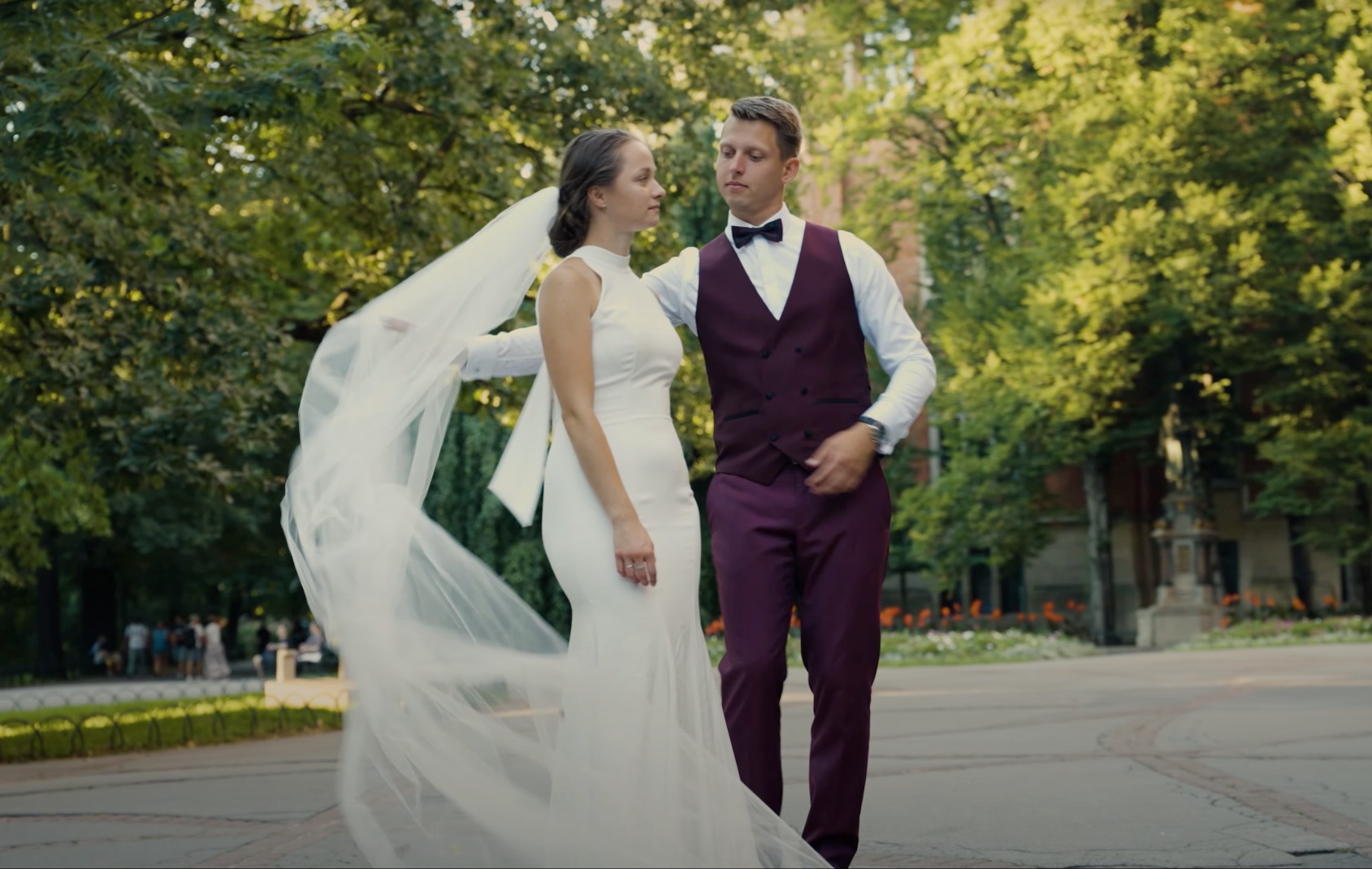 Para młoda spacerująca w parku, panna młoda w prostej białej sukni i długim welonie, pan młody w bordowej kamizelce i muszce.