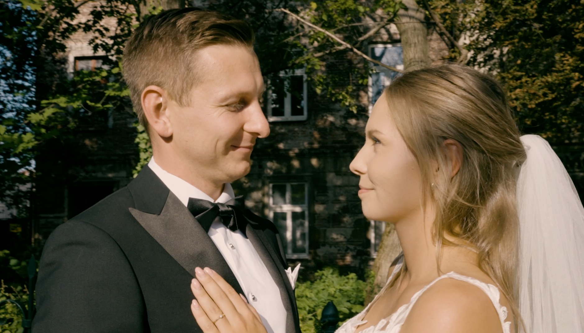 Uśmiechnięta para młoda w zbliżeniu, panna młoda w białej sukni i welonie, pan młody w czarnym smokingu z muszką, na tle budynku z cegły i zieleni.