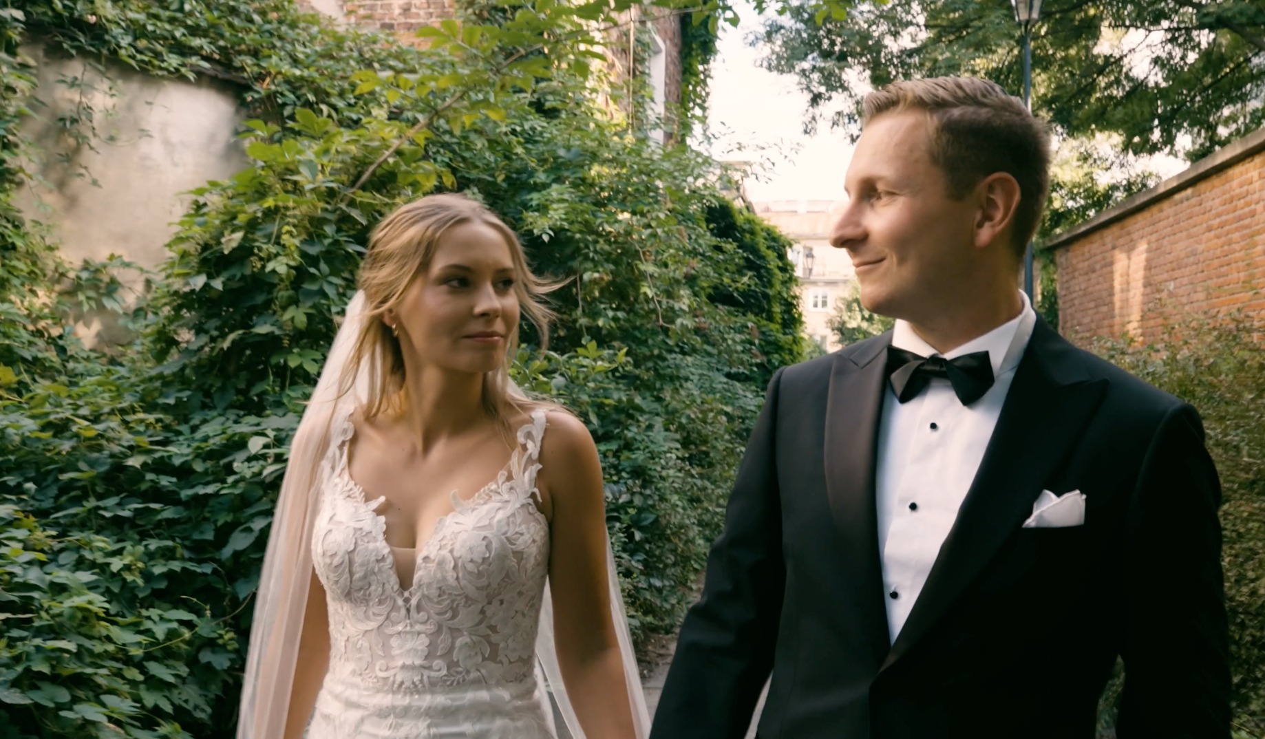 Para młoda spacerująca w parku, panna młoda w koronkowej sukni z welonem, pan młody w smokingu z muchą, w tle zieleń i ceglany mur.