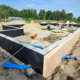 Fasadecraft - Fundamenty domu w trakcie budowy: izolacja przeciwwilgociowa, ocieplenie, rury drenażowe. Widoczny teren budowy i zieleń w tle. Prace ziemne i przygotowanie do dalszych etapów.