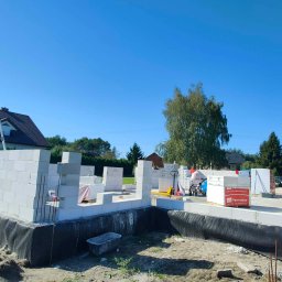 Fasadecraft - Budowa domu jednorodzinnego w trakcie wznoszenia ścian z betonu komórkowego. Widoczne fundamenty, izolacja, palety z bloczkami Termobet i pracownik w oddali.
