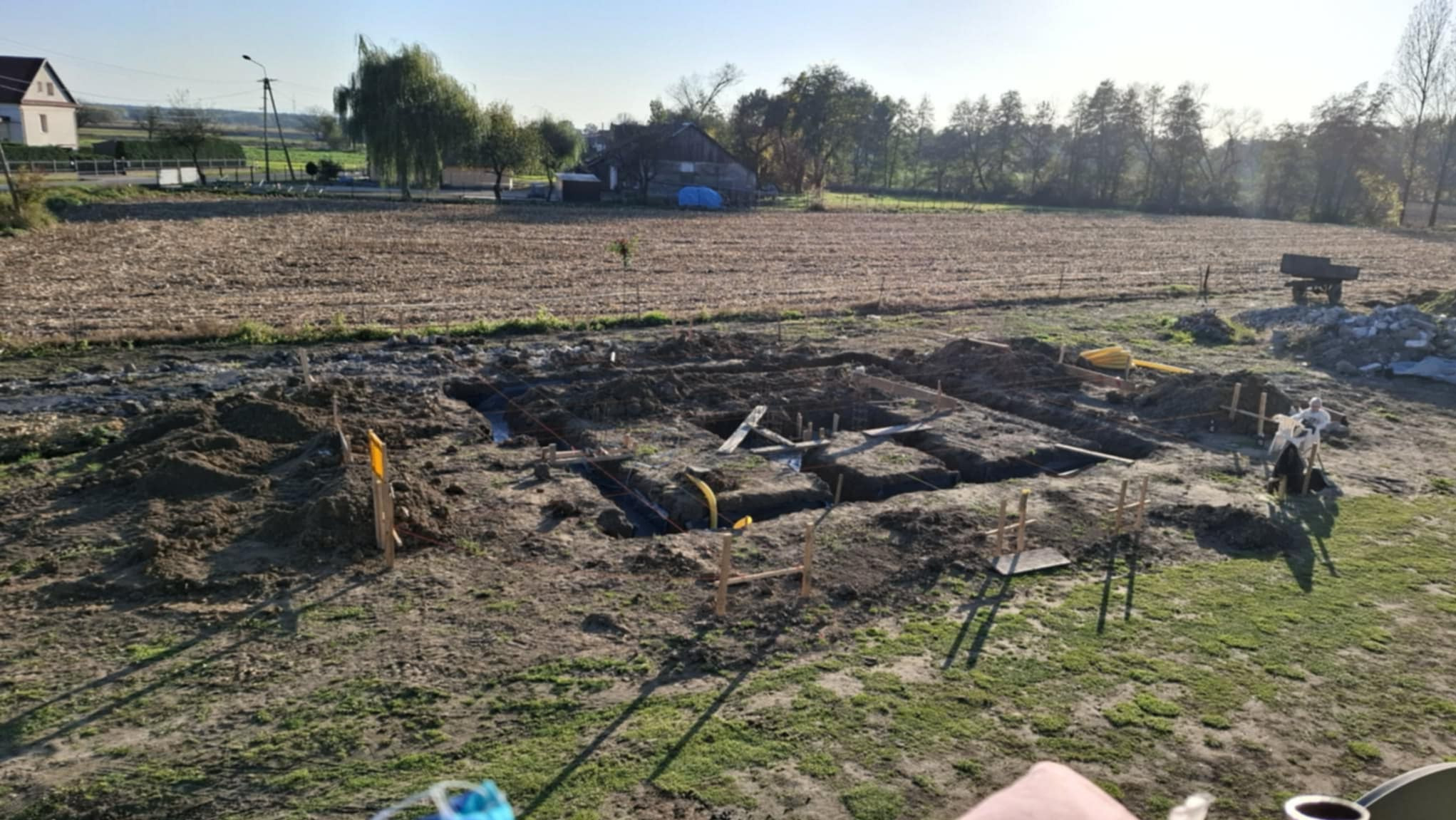 Fundamenty domu w trakcie budowy, widoczne wykopy i szalunki, teren budowy z ziemią i trawą, w tle zabudowania wiejskie i drzewa, słoneczny dzień.