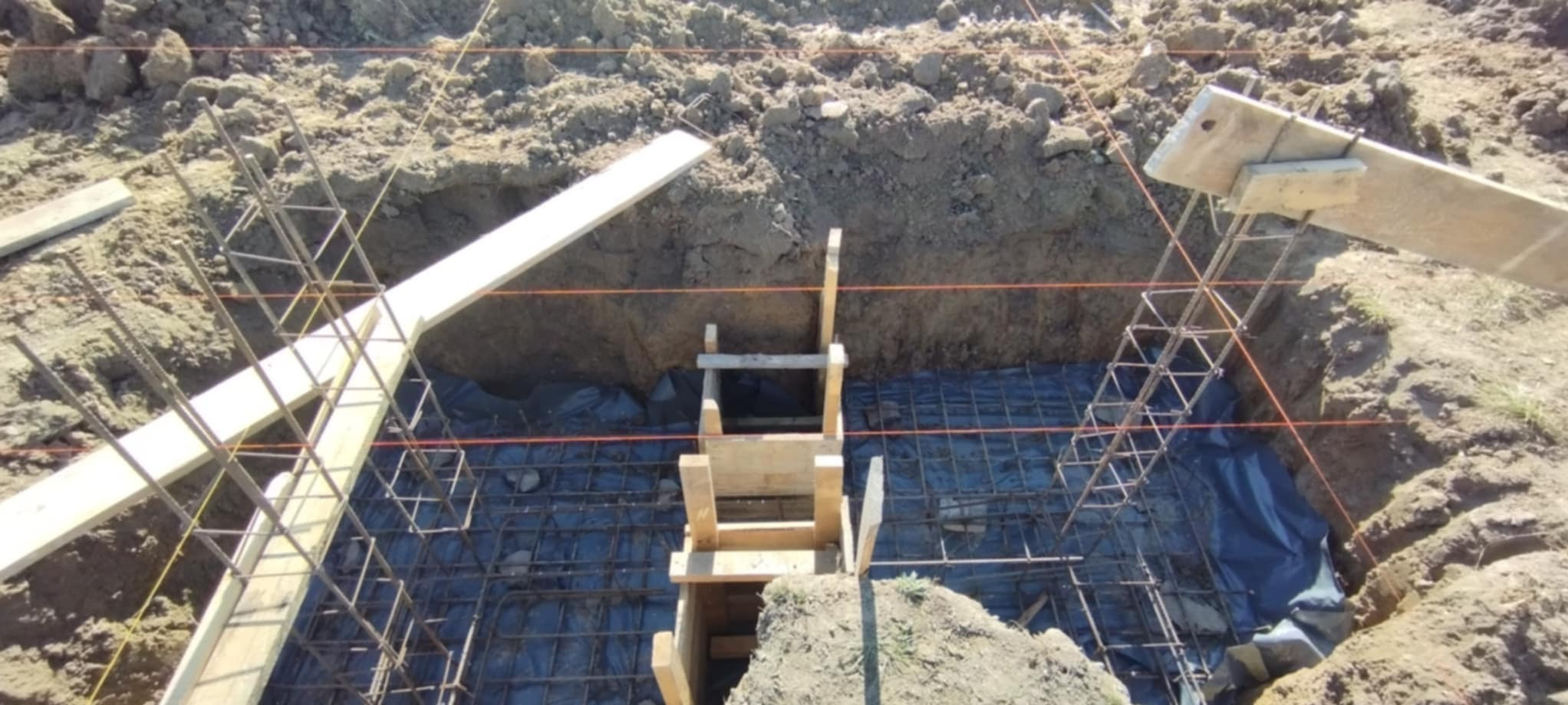 Wykonane wykopy pod fundamenty z ułożonym zbrojeniem i szalunkiem. Widoczne zabezpieczenie folią. Prace ziemne w toku, przygotowanie do wylania betonu.