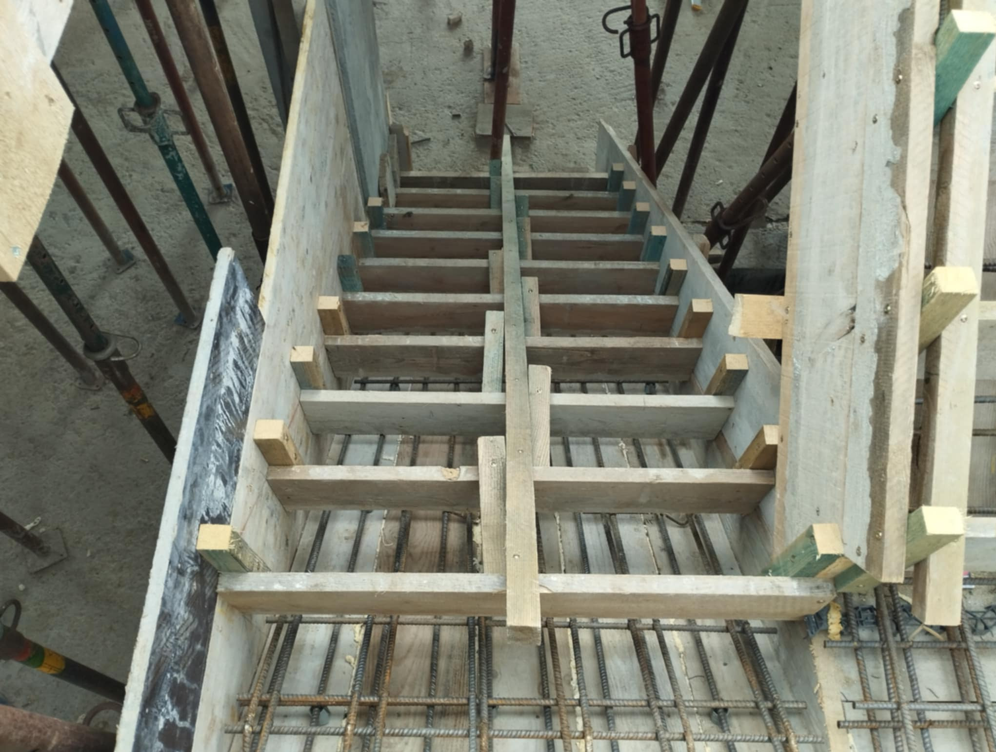 Szczegółowe ujęcie szalunku schodów z widocznym zbrojeniem, przygotowanego do zalania betonem. Widok z góry na drewnianą konstrukcję i stalowe pręty.