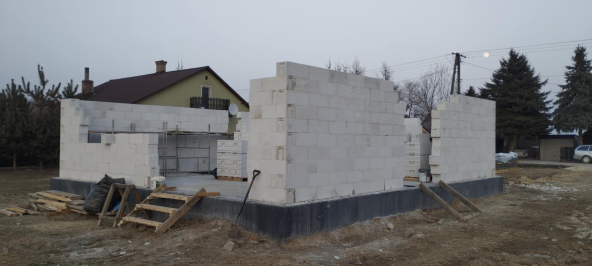 Wznoszenie ścian parteru domu z białych bloczków, widoczne rusztowania i materiały budowlane. W tle dom z brązowym dachem i drzewa.