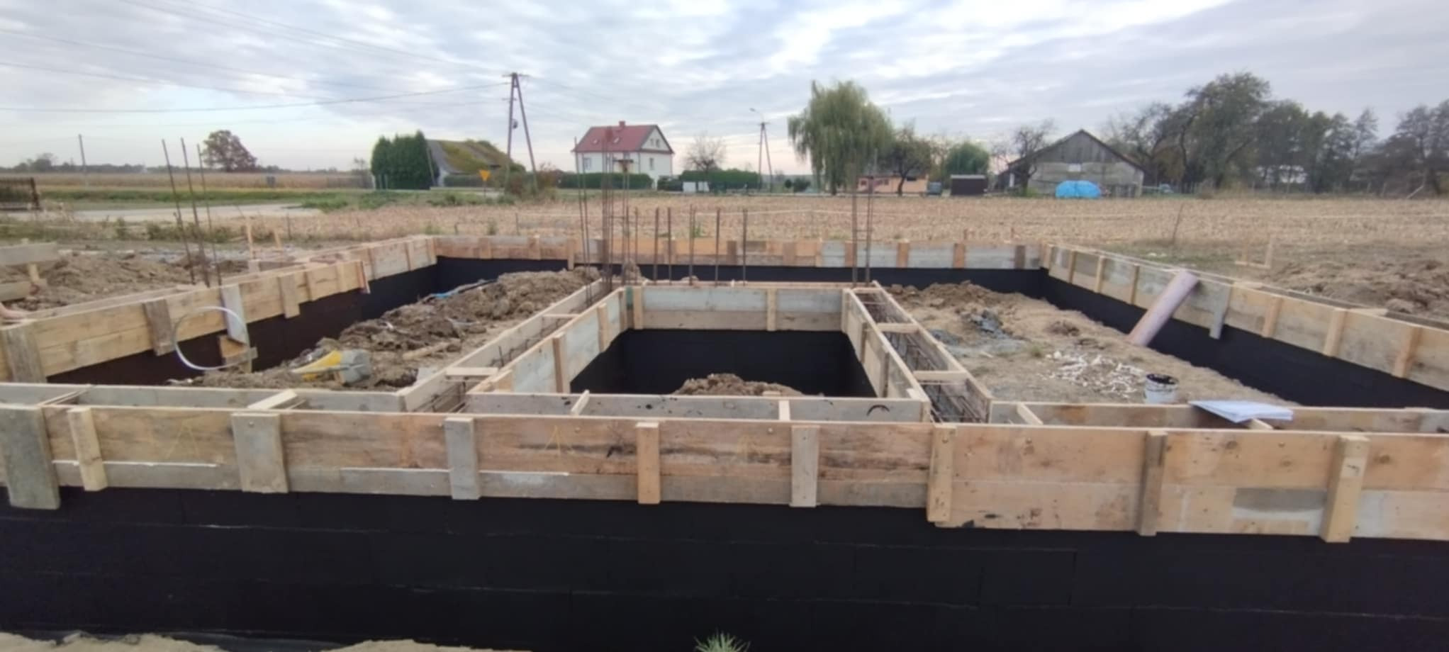 Fundamenty domu w trakcie budowy: drewniane szalunki, izolacja przeciwwilgociowa, zbrojenie. W tle krajobraz wiejski z domami i polami.