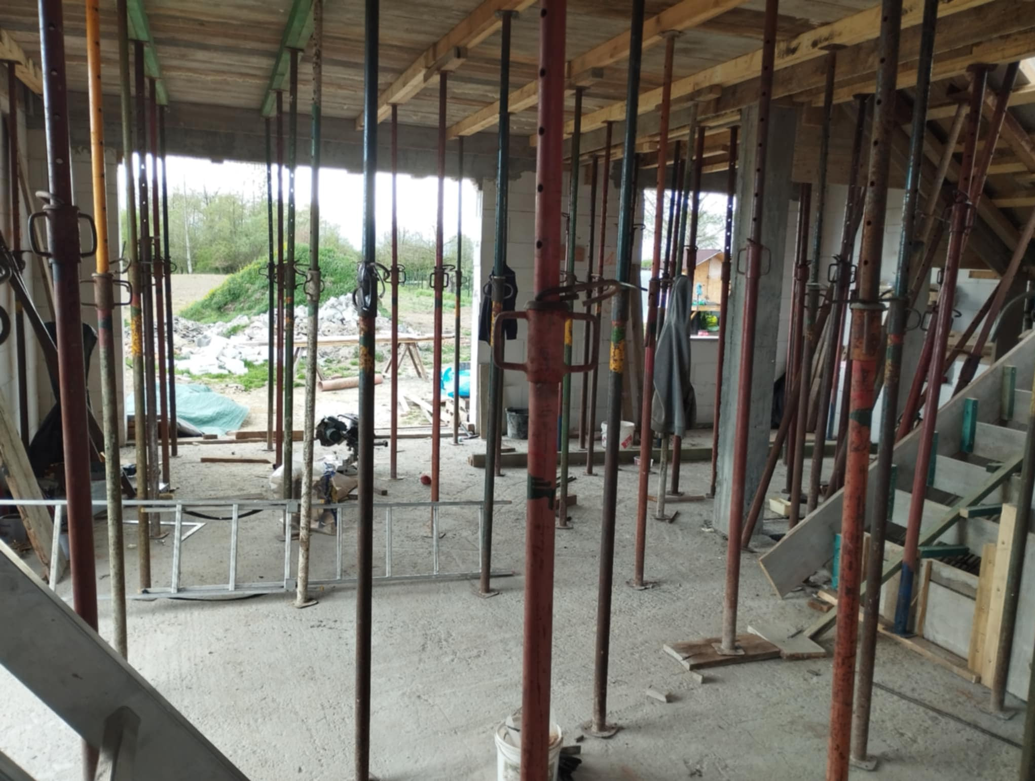 Wnętrze w trakcie budowy z licznymi metalowymi stemplami podtrzymującymi strop, widoczne elementy szalunków i drabina. Surowy stan, betonowa podłoga.