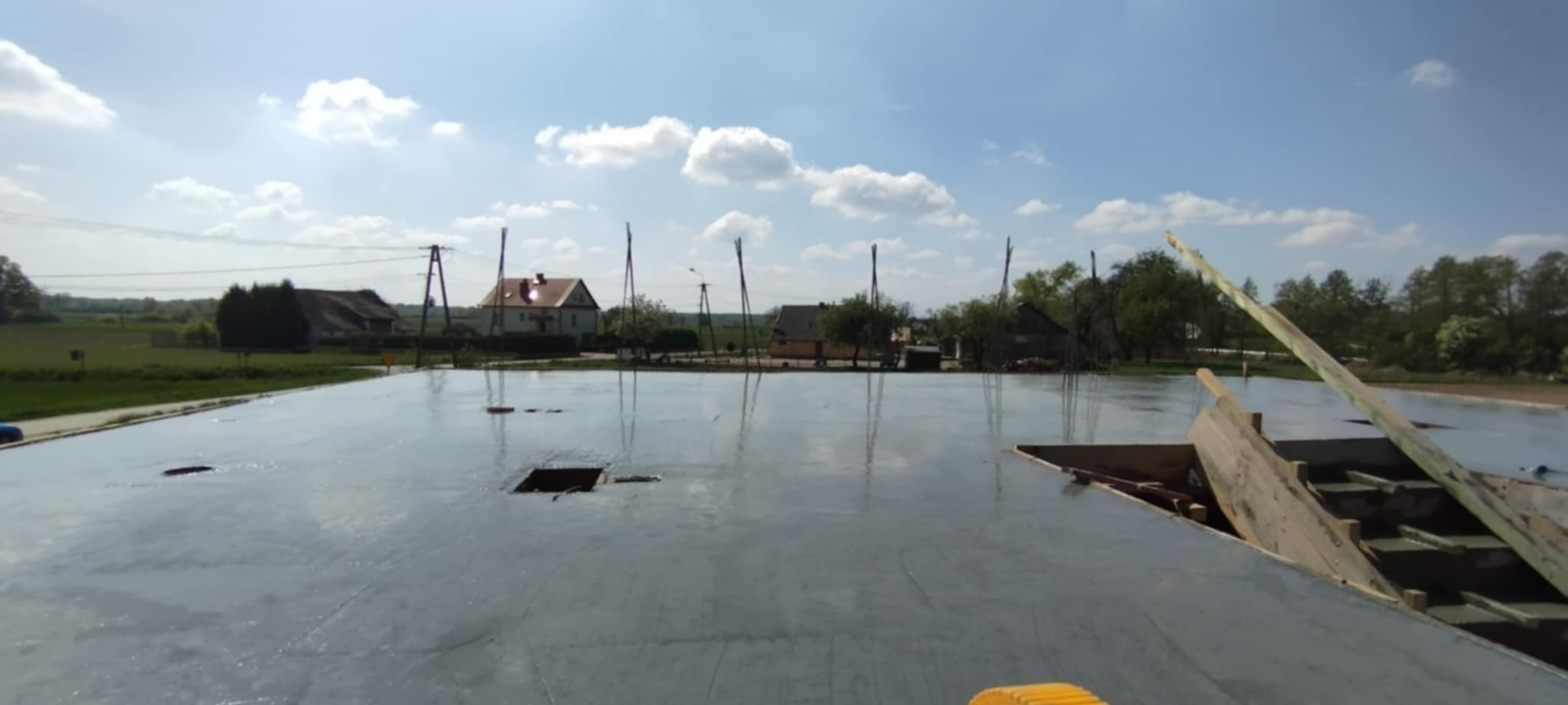 Świeżo wylana, gładka posadzka betonowa na zewnątrz budynku, z widocznymi elementami szalunku i odbiciami nieba. W tle zabudowania wiejskie i słupy energetyczne.