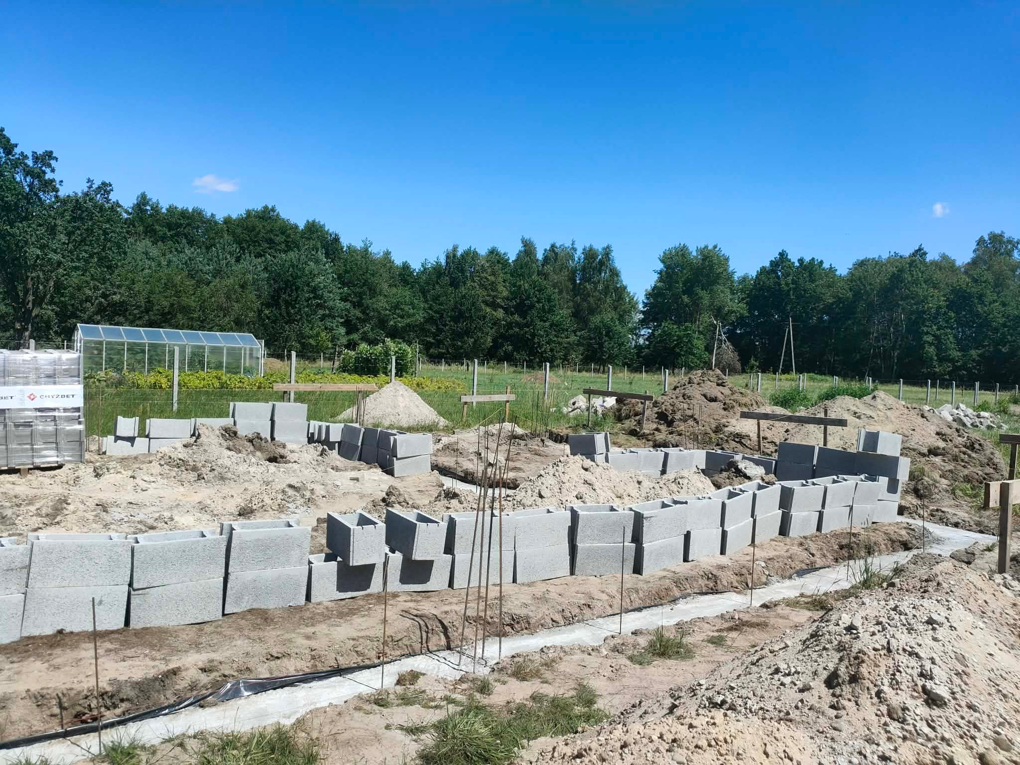Fundamenty z bloczków betonowych na budowie domu jednorodzinnego, widok na teren z zielenią i błękitnym niebem w tle, w słoneczny dzień.