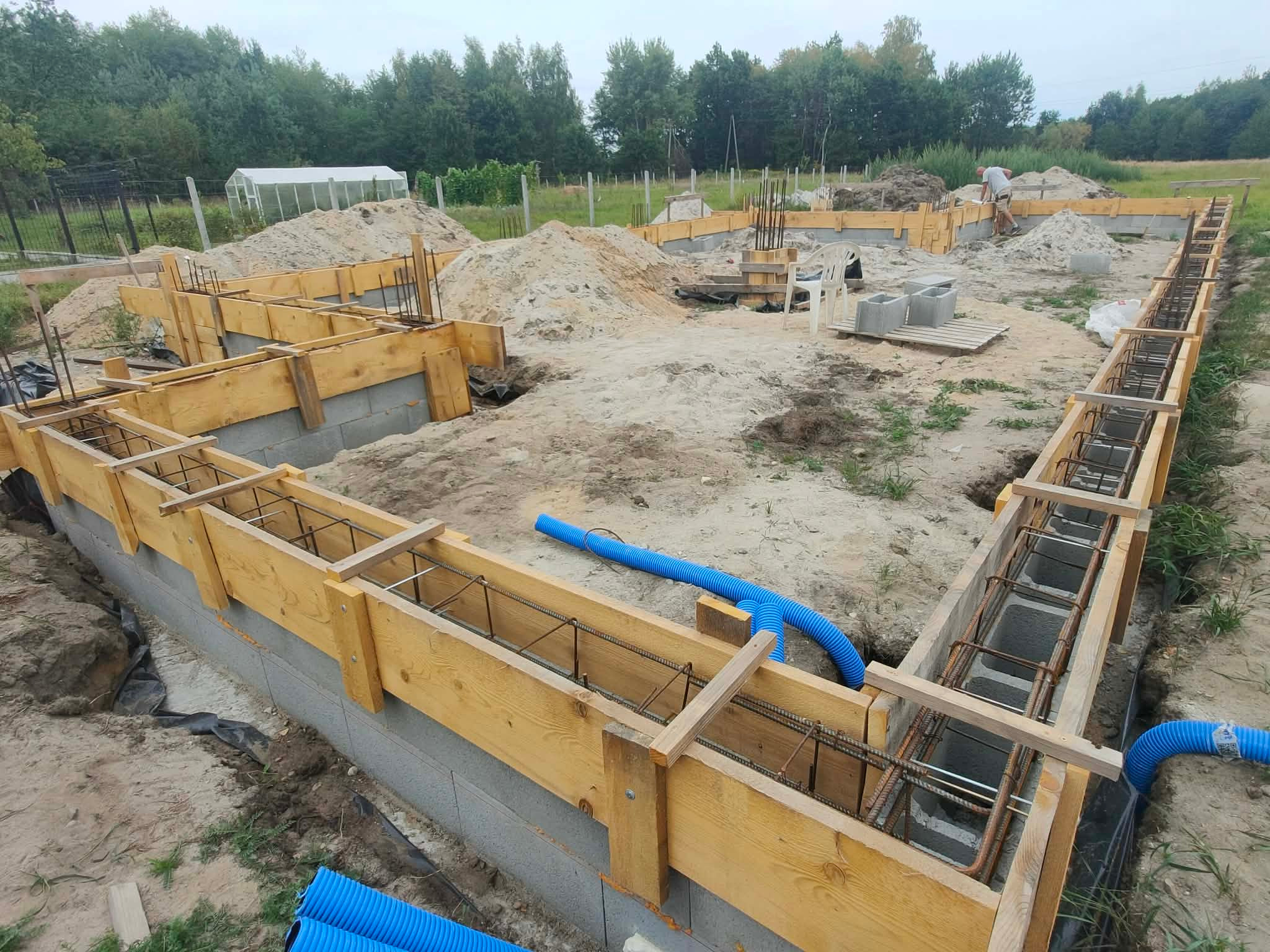 Fundamenty domu w trakcie budowy: szalunki z drewna, zbrojenie stalowe, niebieskie rury drenażowe. W tle widoczny teren budowy i zieleń.