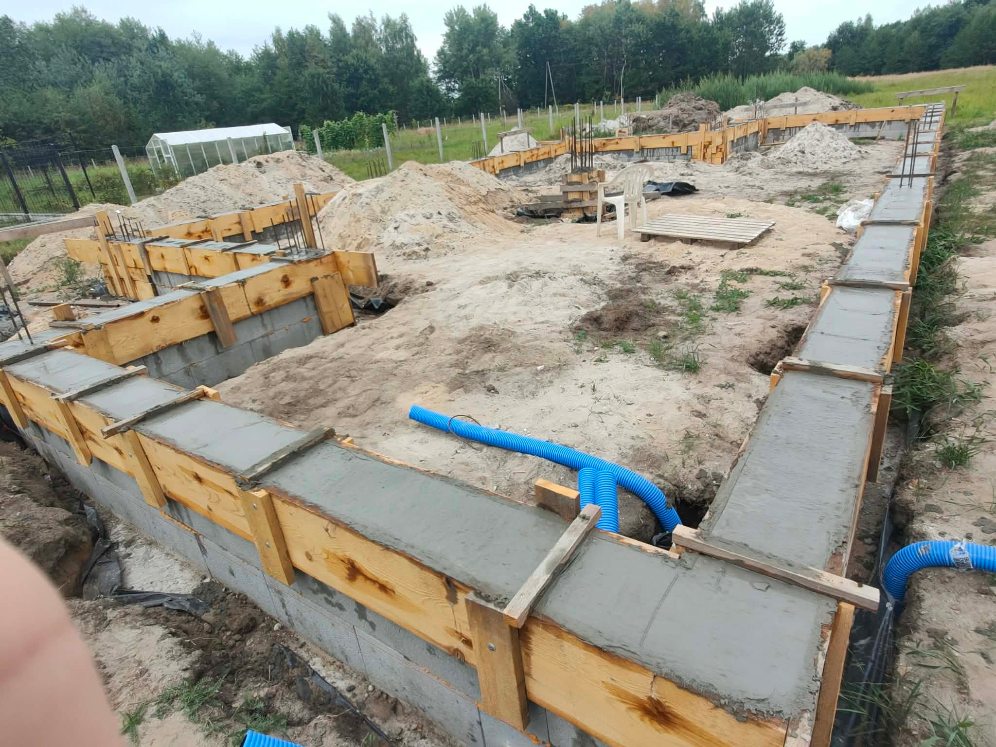 Fundamenty domu w trakcie budowy: betonowe ściany szalunkowe, wystające pręty zbrojeniowe, niebieskie rury drenażowe, ziemia i piasek. Widok z góry na plac budowy.