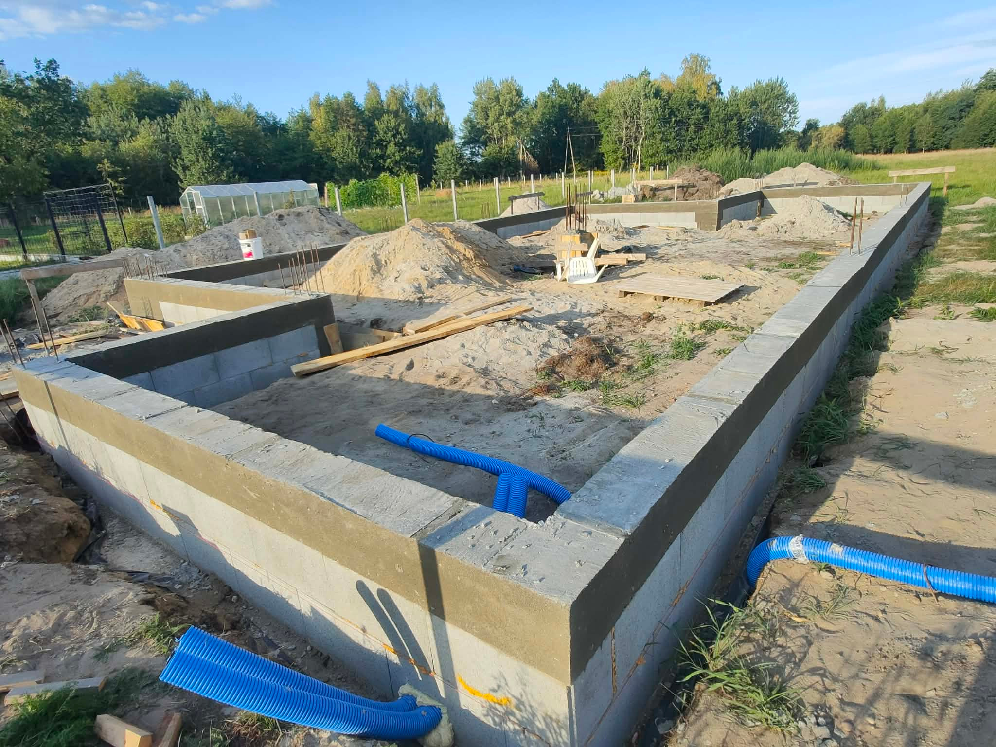 Fundamenty domu jednorodzinnego w trakcie budowy, z widocznymi elementami izolacji i niebieskimi rurami drenażowymi. W tle zieleń drzew i częściowo widoczna szklarnia.