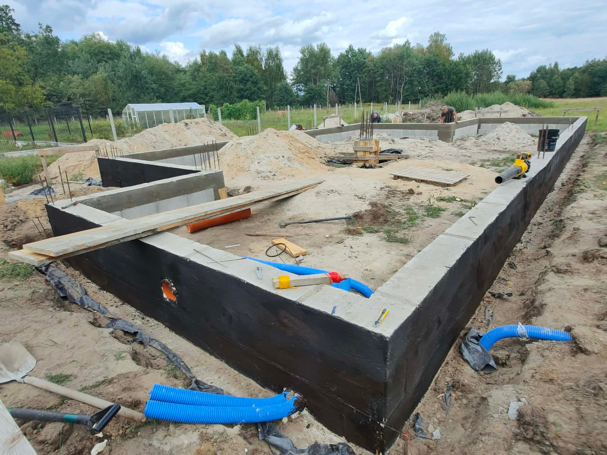 Fundamenty domu w trakcie budowy, z widoczną izolacją przeciwwilgociową, rurami instalacyjnymi i narzędziami na placu budowy. W tle zieleń i szklarnia.