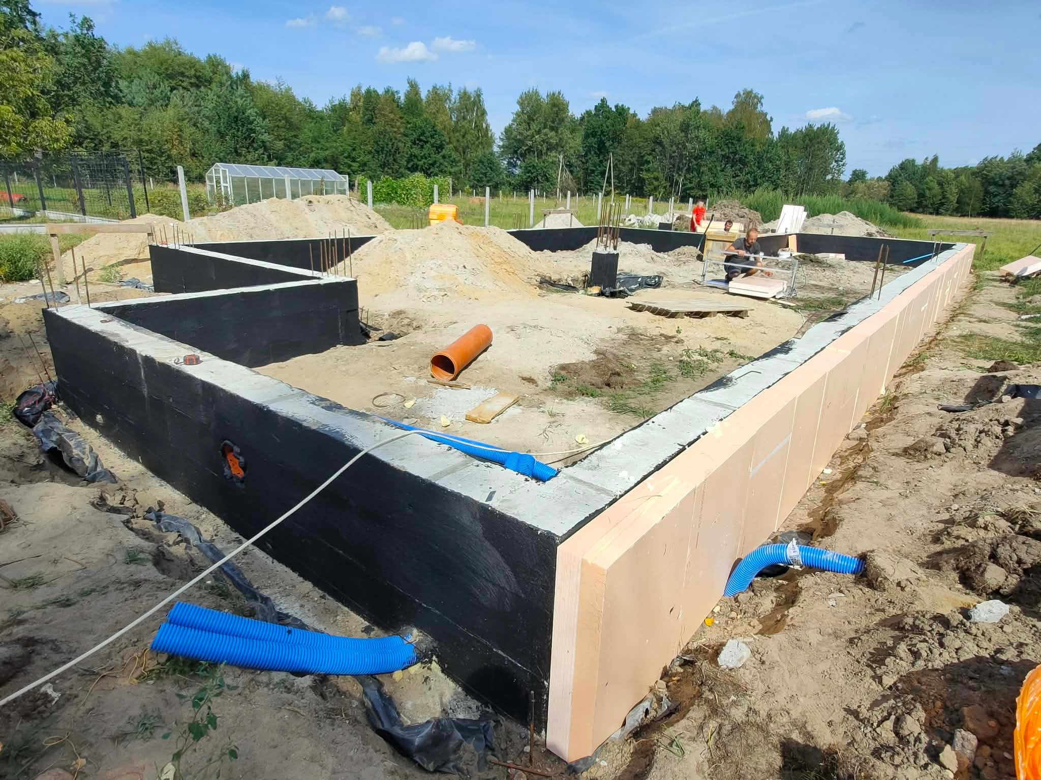 Fundamenty domu w trakcie budowy: izolacja przeciwwilgociowa, ocieplenie, rury drenażowe. Widoczny teren budowy i zieleń w tle. Prace ziemne i przygotowanie do dalszych etapów.