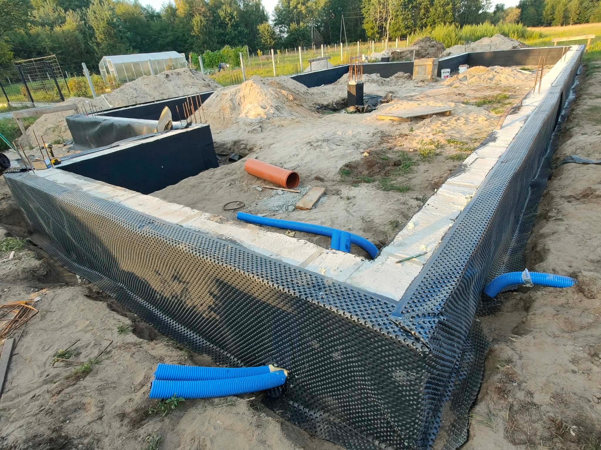 Fundamenty domu w trakcie budowy: izolacja pionowa z folii kubełkowej, drenaż z niebieskich rur karbowanych, zbrojenie pionowe. Widok z góry na plac budowy.