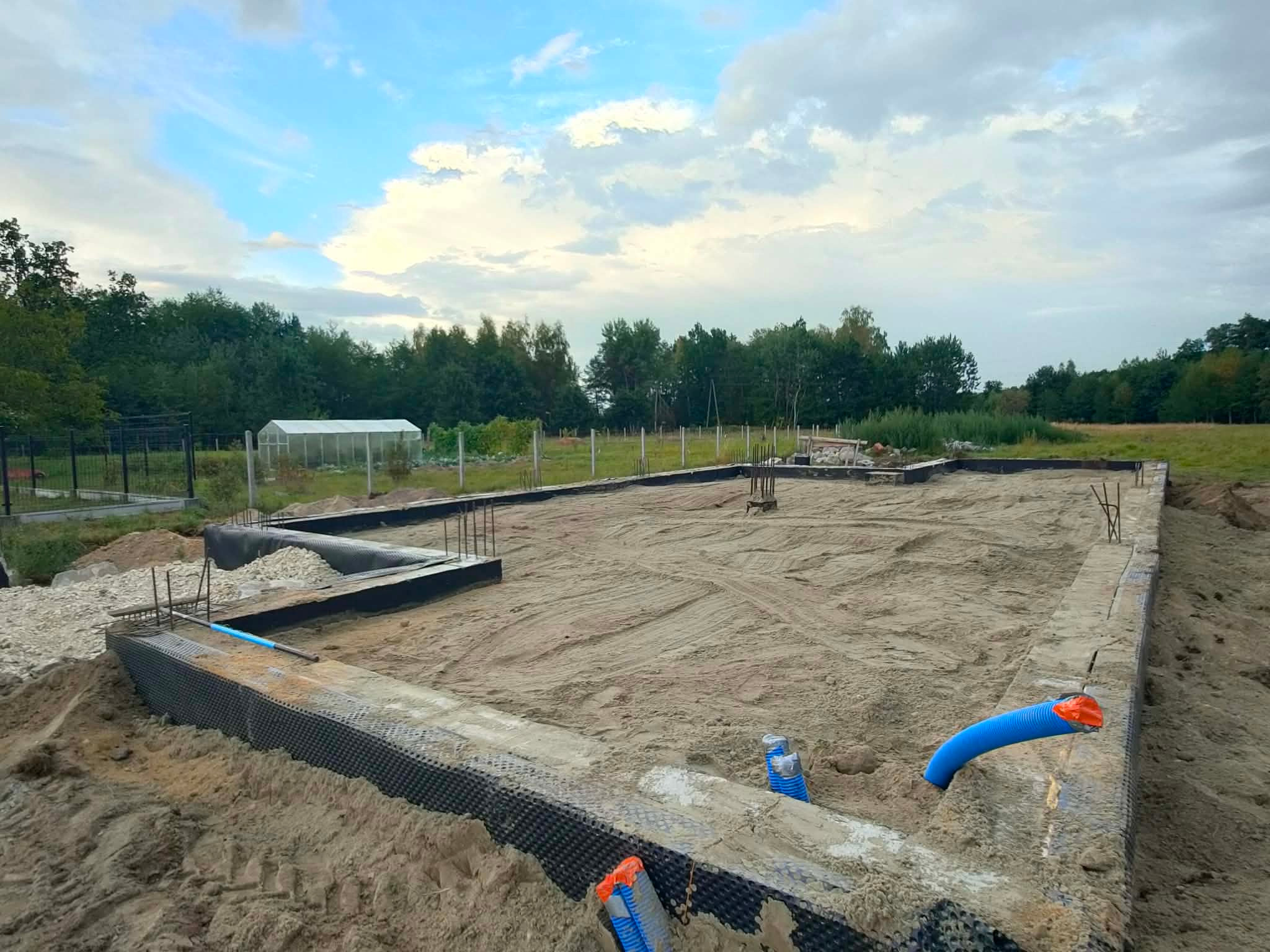 Fundamenty domu jednorodzinnego w Brzesku, z widoczną hydroizolacją i instalacją kanalizacyjną. W tle zieleń i szklarnia, niebo z chmurami.