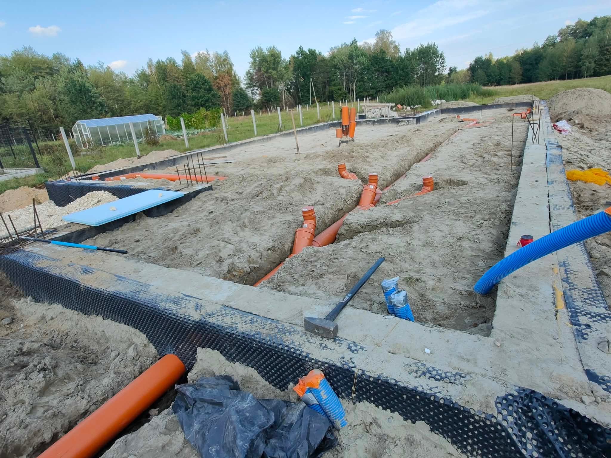 Fundamenty domu w trakcie budowy: zbrojenie, izolacja, rury kanalizacyjne w wykopach. Widoczne narzędzia i otoczenie placu budowy.