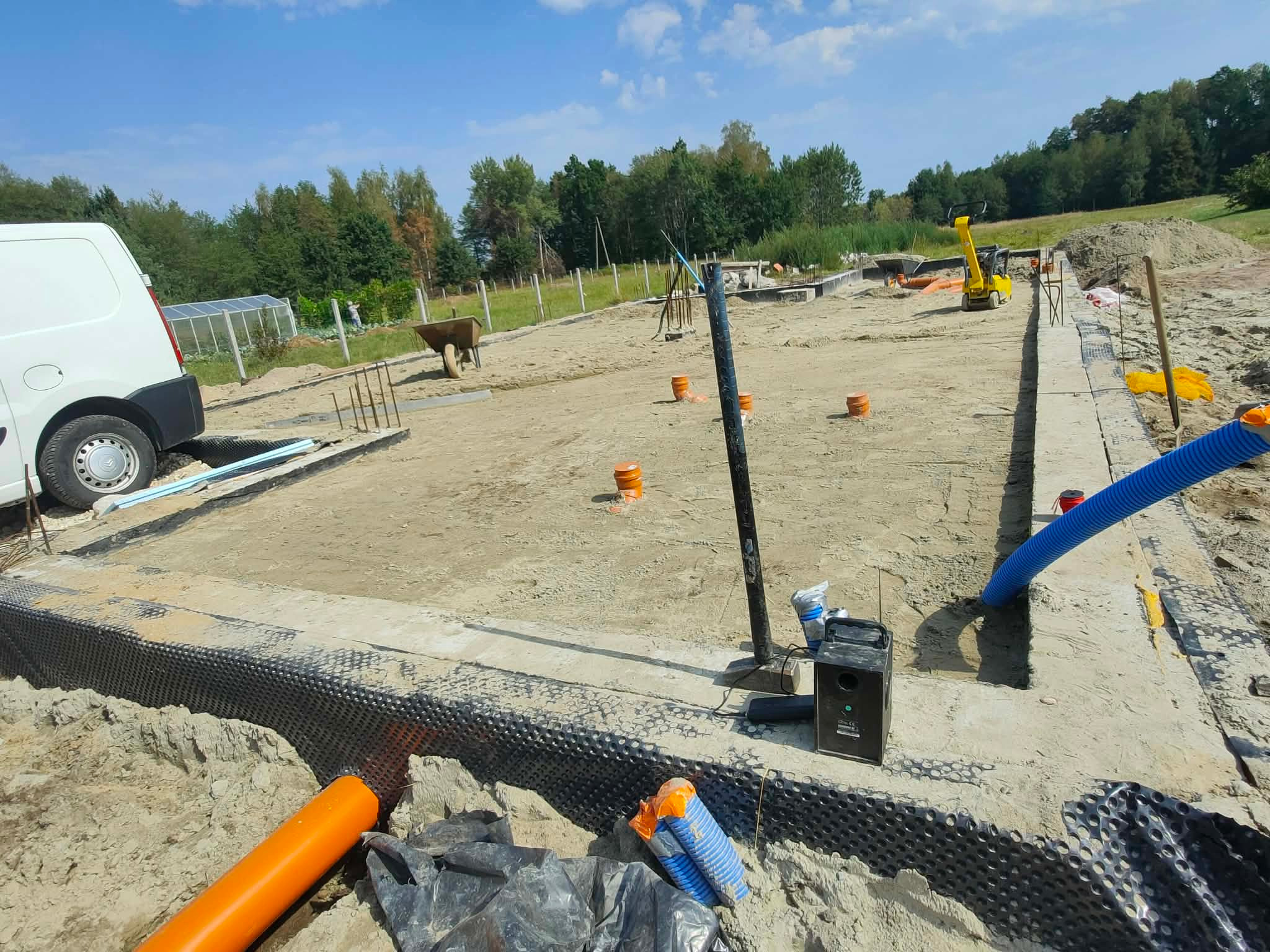 Fundamenty domu w trakcie budowy, widoczne izolacje, rury drenażowe, zagęszczarka i częściowo biały bus. W tle zieleń i błękitne niebo.