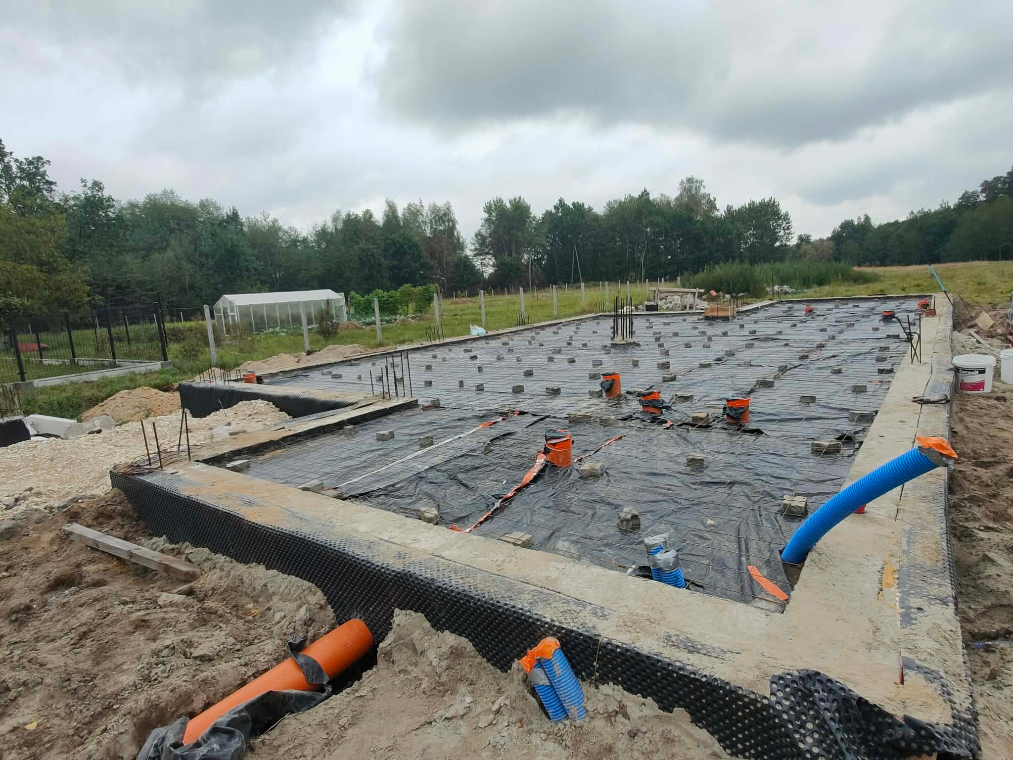 Fundamenty domu w trakcie budowy: zbrojenie, izolacja, rury drenażowe. Widok na teren budowy z zielenią w tle i pochmurnym niebem.