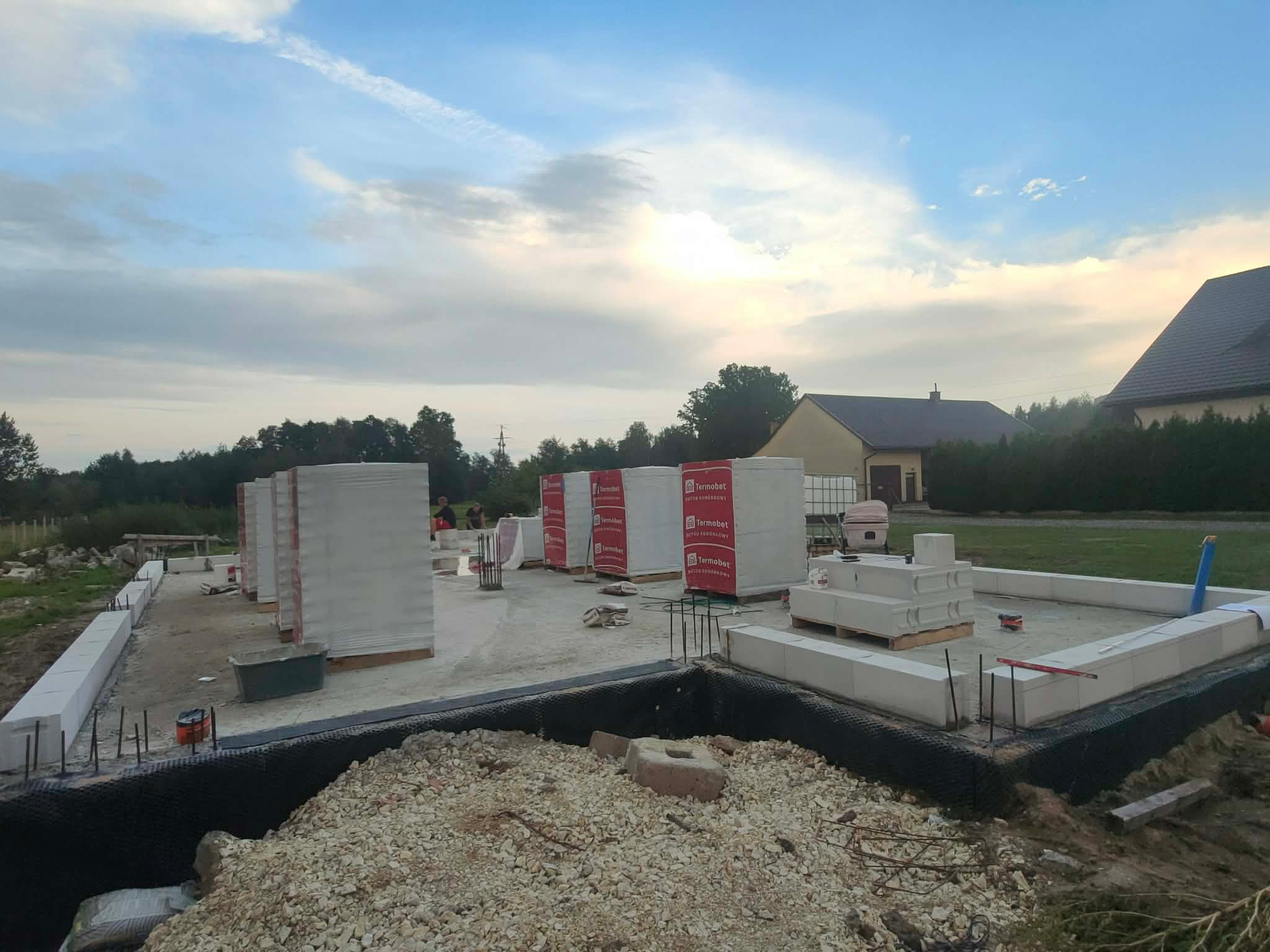 Budowa domu jednorodzinnego: fundamenty zbrojone, bloczki Termobet, widok na plac budowy w trakcie wznoszenia ścian. Niebo z chmurami w tle.