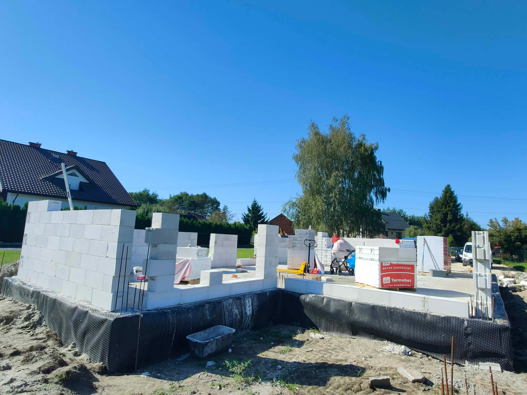 Budowa domu jednorodzinnego w trakcie wznoszenia ścian z betonu komórkowego. Widoczne fundamenty, izolacja, palety z bloczkami Termobet i pracownik w oddali.