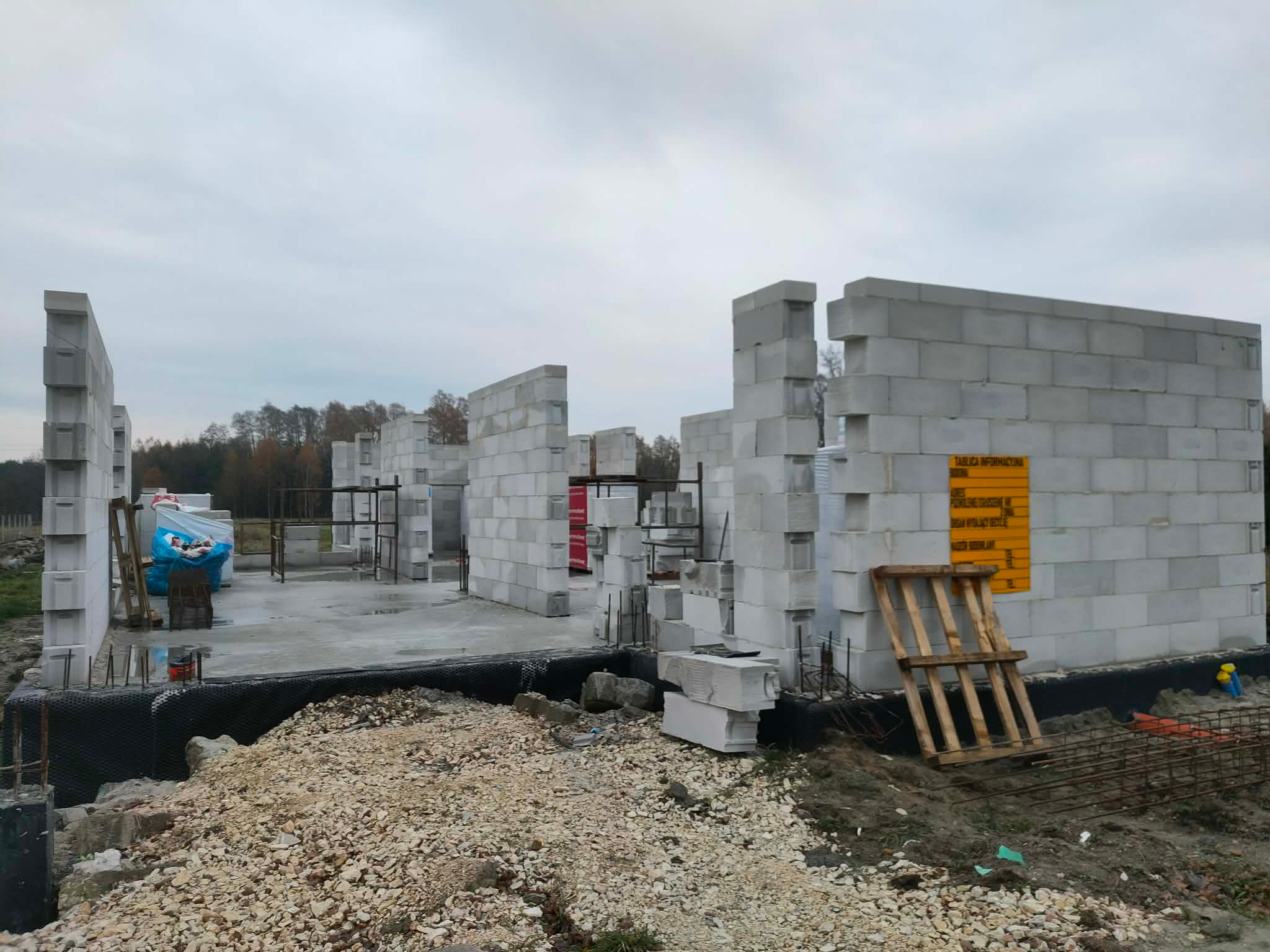 Wznoszenie ścian domu z bloczków betonowych. Widoczna hydroizolacja fundamentów, paleta oparta o ścianę i tablica informacyjna. Surowy stan budowy.