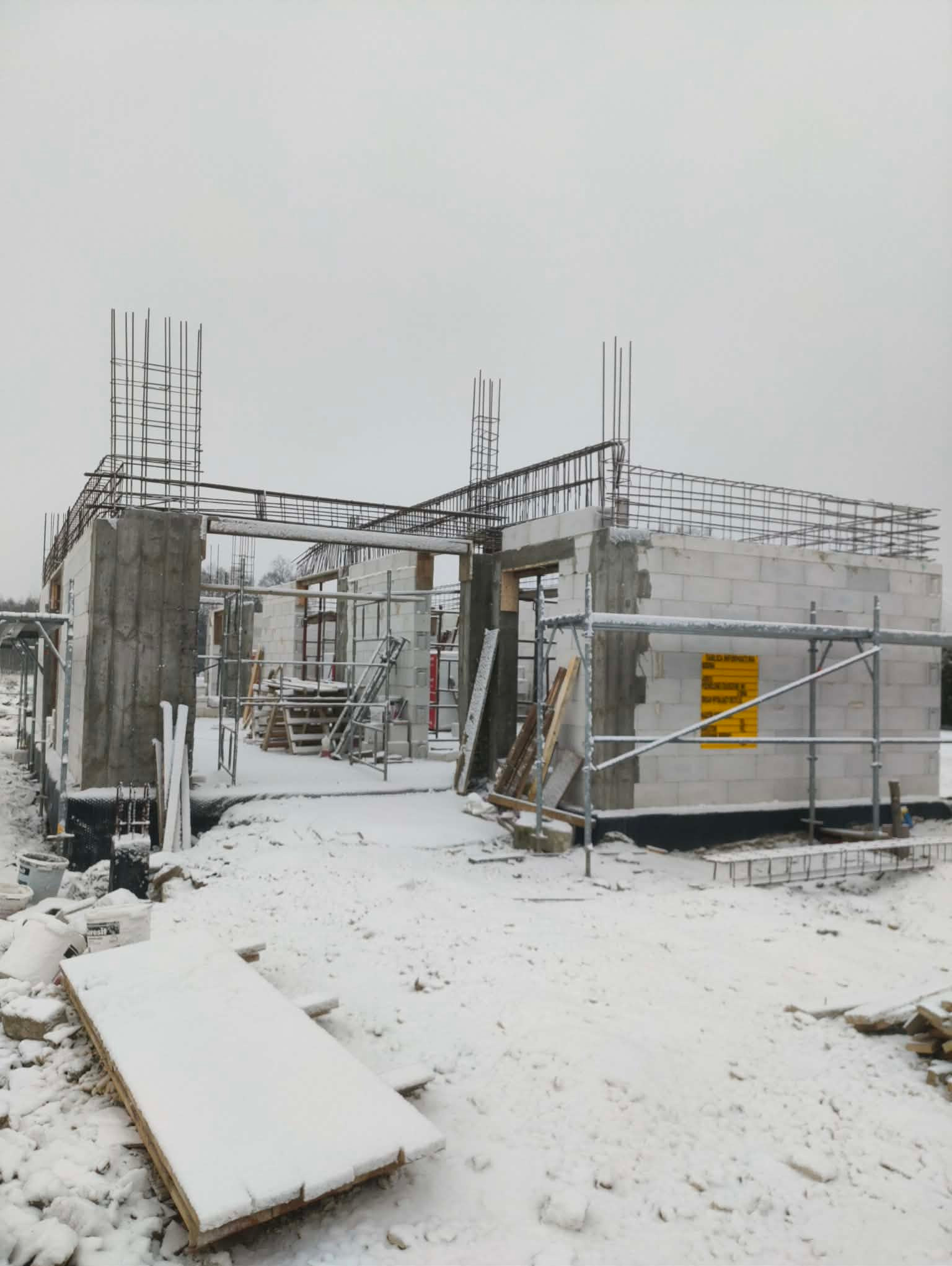Surowy stan domu w zimowej scenerii: ściany z betonu komórkowego, zbrojenia, rusztowania, śnieg. Widok na budowę w trakcie wznoszenia, w pochmurny dzień.