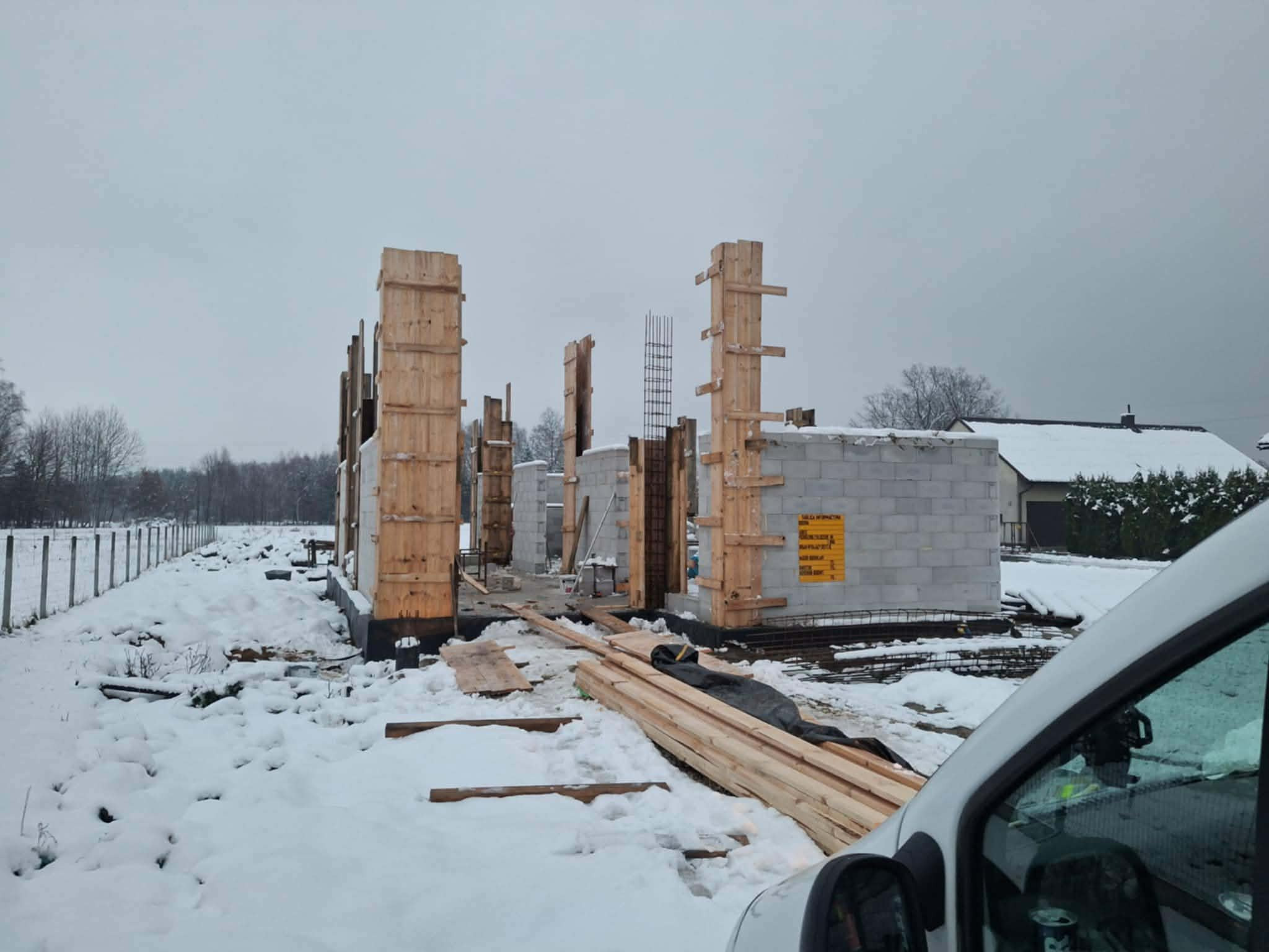 Budowa domu w zimowej scenerii: drewniane szalunki kolumn i fragment muru z bloczków betonowych, pokryte śniegiem. Widoczne materiały budowlane i zbrojenia.