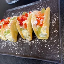 Food Miód i Orzeszki - Trzy mini desery w kształcie taco na czarnym talerzu, wypełnione kremem i udekorowane truskawkami i zielonymi winogronami, posypane cukrem pudrem.