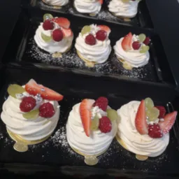 Trzy rzędy mini bez z kremem i owocami (truskawki, maliny, winogrona) posypane cukrem pudrem i srebrnymi kuleczkami, ułożone na czarnych prostokątnych tacach.