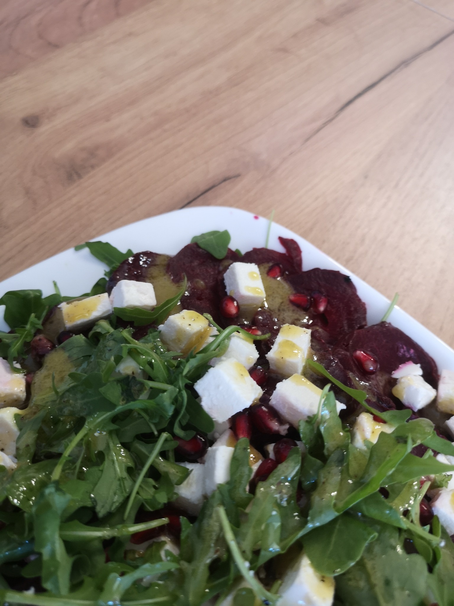 Sałatka z buraków, rukoli, sera feta i granatu, polana sosem, podana na białym talerzu na drewnianym stole.