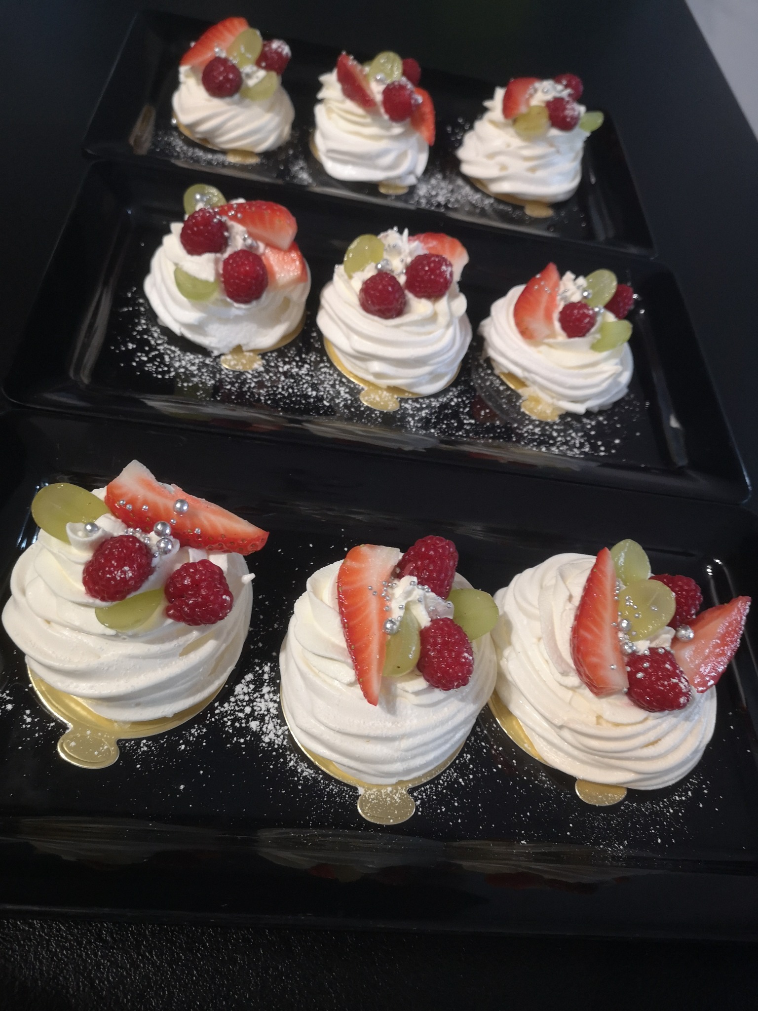 Trzy rzędy mini bez z kremem i owocami (truskawki, maliny, winogrona) posypane cukrem pudrem i srebrnymi kuleczkami, ułożone na czarnych prostokątnych tacach.