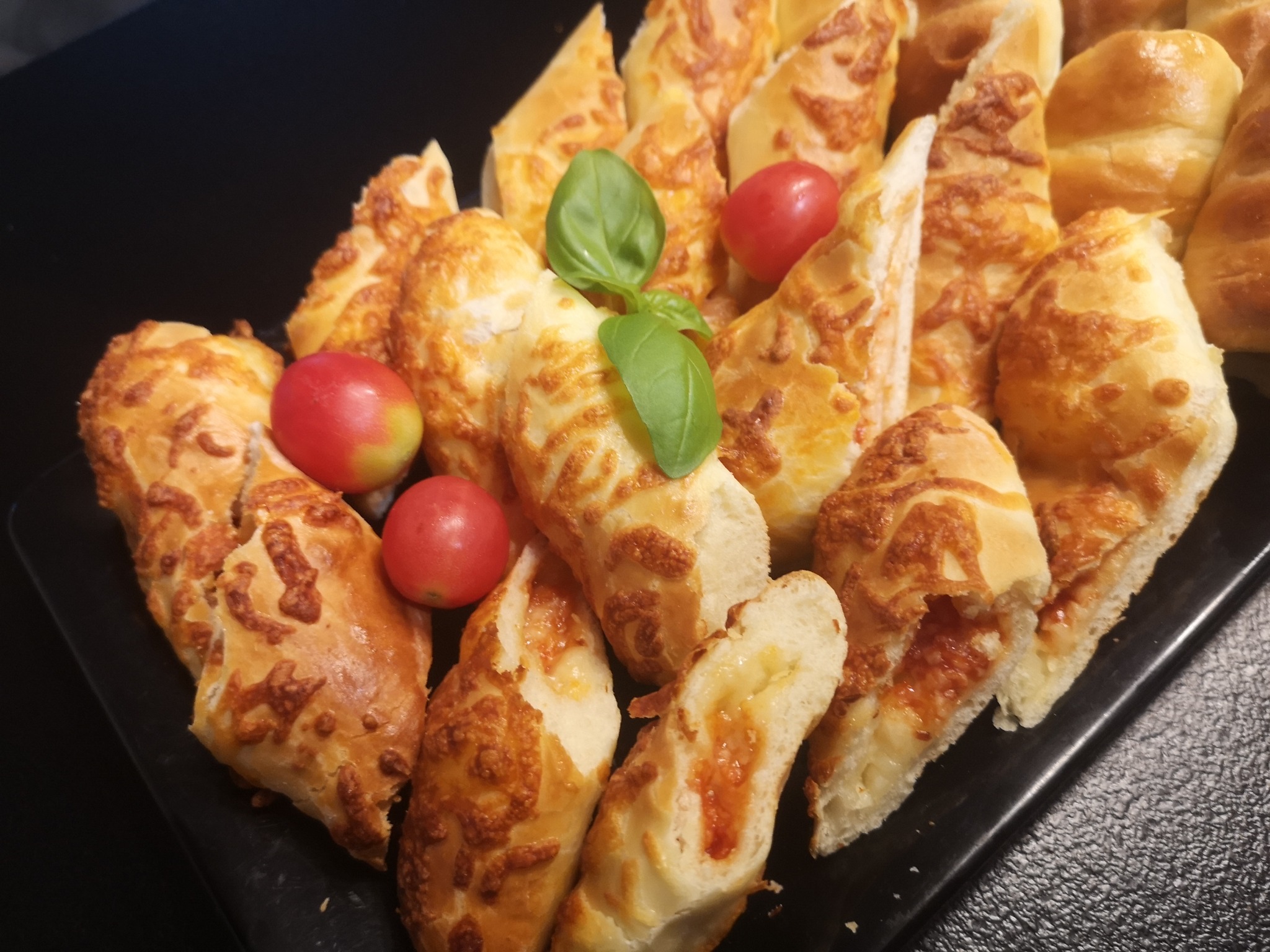 Kawałki pieczywa z serem i pomidorami na czarnym talerzu, udekorowane listkami bazylii i pomidorkami koktajlowymi.