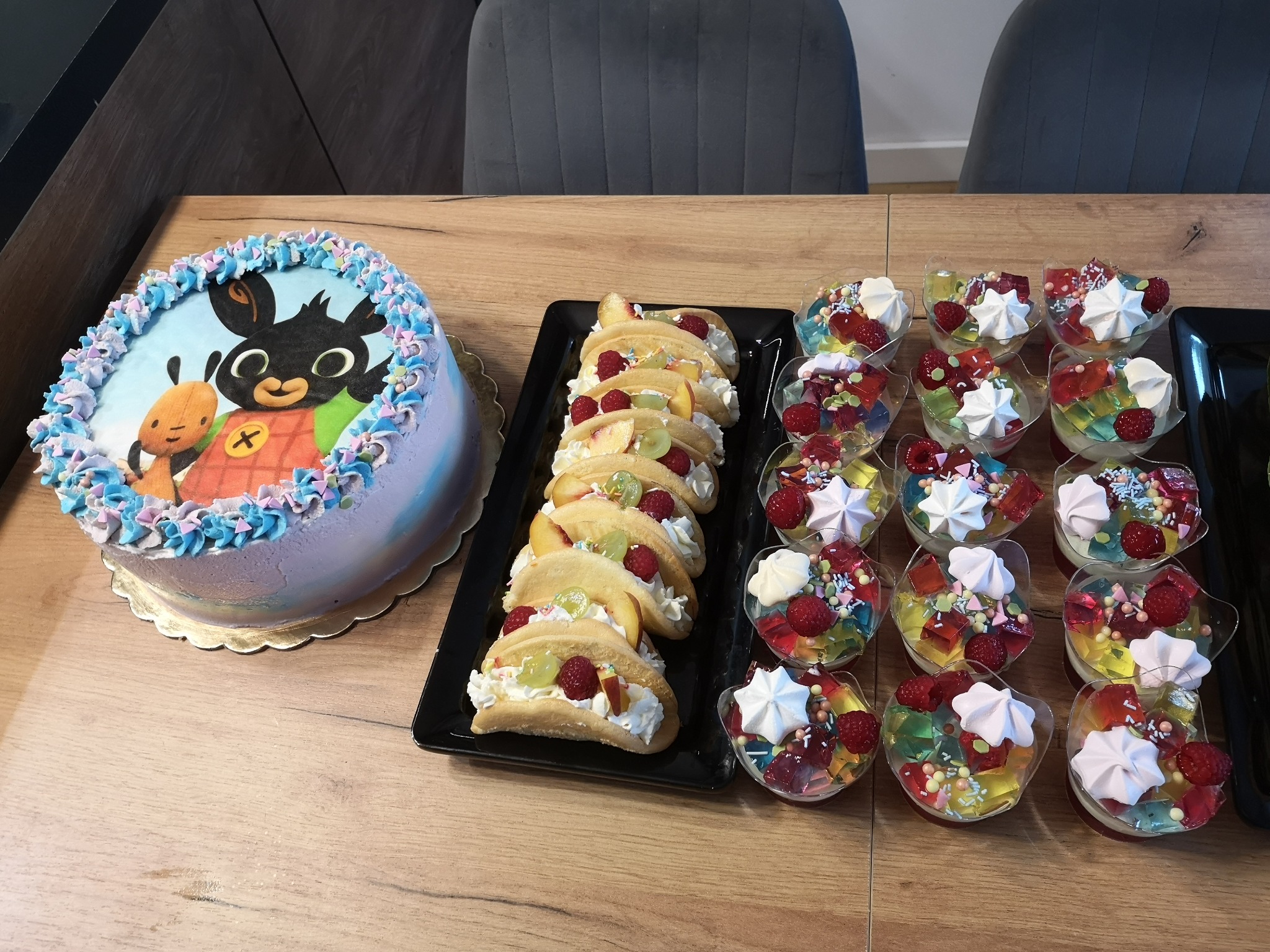 Urodzinowy tort z postacią z bajki Bing, desery w kształcie tacos z owocami i bitej śmietany oraz galaretki z owocami i bezami w plastikowych pucharkach na drewnianym stole.