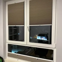 Zamknięte, beżowe osłony okienne plisowane w białym oknie PCV, widok na nocne miasto odbijający się w szybach.