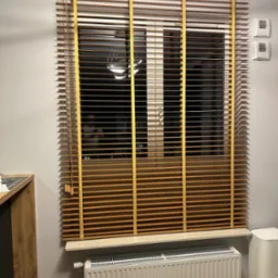 Zamknięte poziome żaluzje w odcieniach brązu z żółtymi taśmami na oknie, widoczny grzejnik pod parapetem i fragment robota sprzątającego.