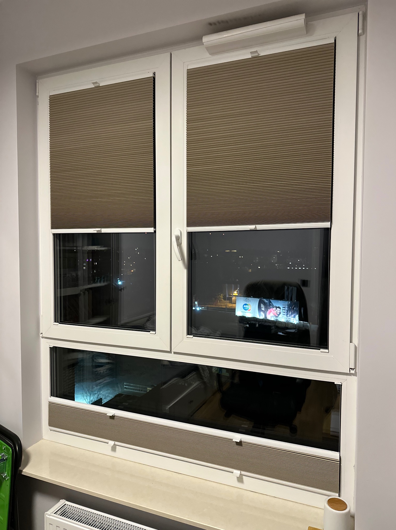 Zamknięte, beżowe osłony okienne plisowane w białym oknie PCV, widok na nocne miasto odbijający się w szybach.