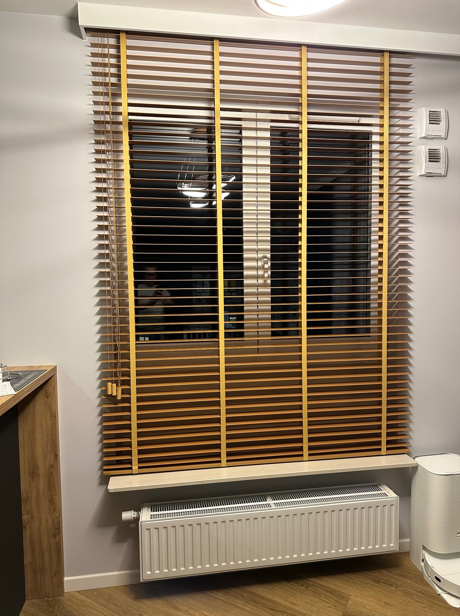 Zamknięte poziome żaluzje w odcieniach brązu z żółtymi taśmami na oknie, widoczny grzejnik pod parapetem i fragment robota sprzątającego.