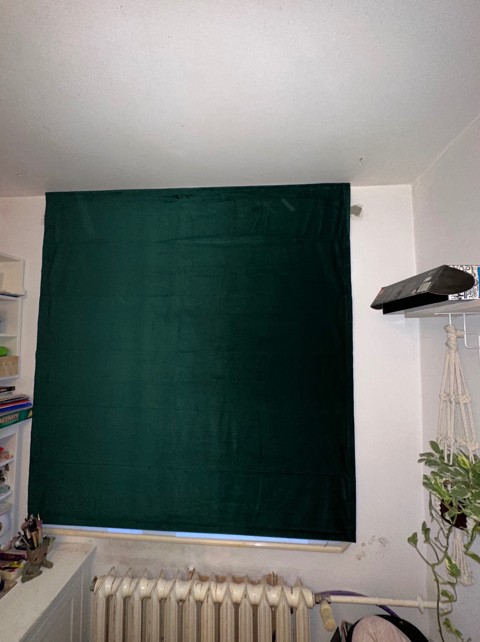 Ciemnozielona, aksamitna zasłona okienna w pokoju z grzejnikiem żeberkowym i półką na ścianie, lekko niedoskonałe wykończenie ścian.
