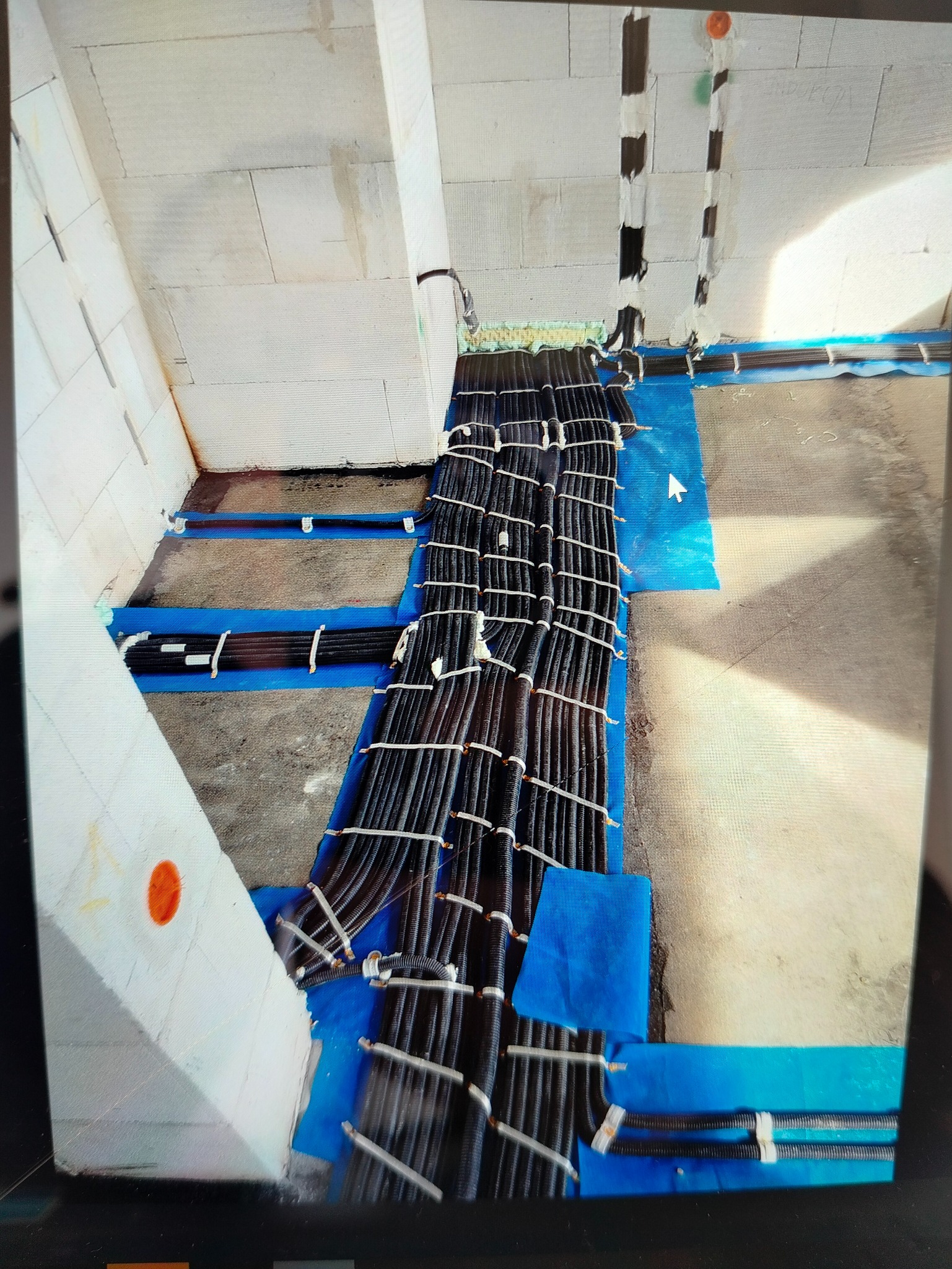 Równolegle ułożone wiązki czarnych rur karbowanych z kablami, zabezpieczone białymi opaskami, prowadzone po podłodze wyłożonej niebieską folią ochronną w surowym pomieszczeniu.