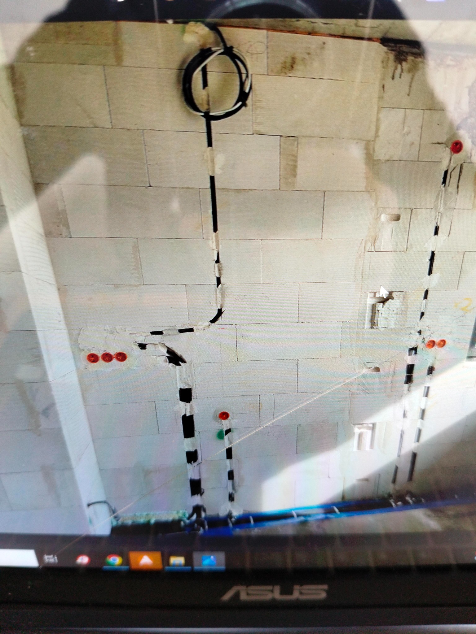 Surowa ściana z betonu komórkowego z poprowadzonymi przewodami elektrycznymi, zabezpieczonymi taśmą, widoczne puszki pod gniazdka, ujęcie z dołu, widoczny fragment ekranu laptopa.