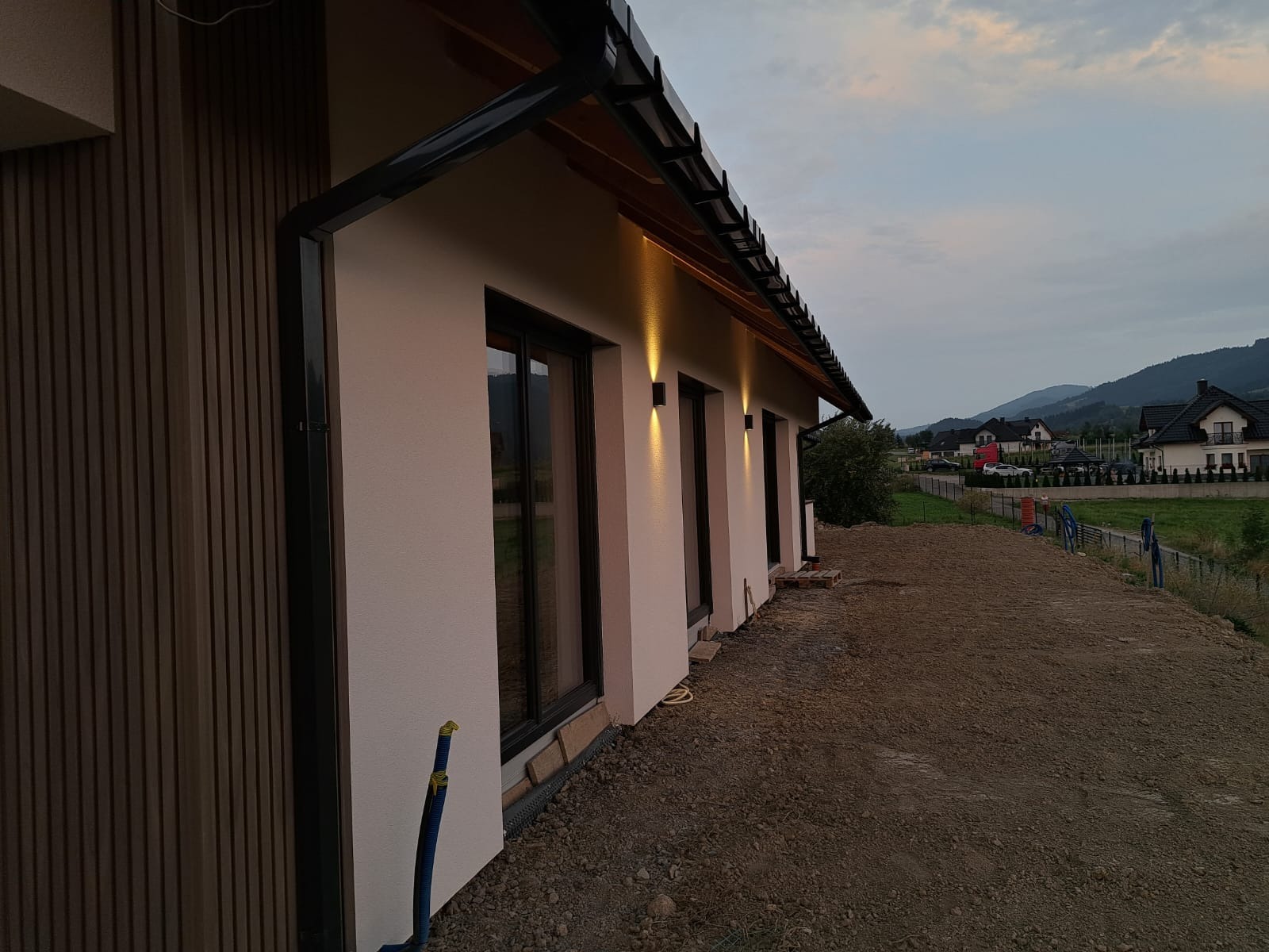 Elewacja domu jednorodzinnego z zamontowanym oświetleniem zewnętrznym, widoczne lampy ścienne emitujące ciepłe światło w górę i w dół, brązowa okładzina pionowa, czarna rynna, tło górzyste, teren...