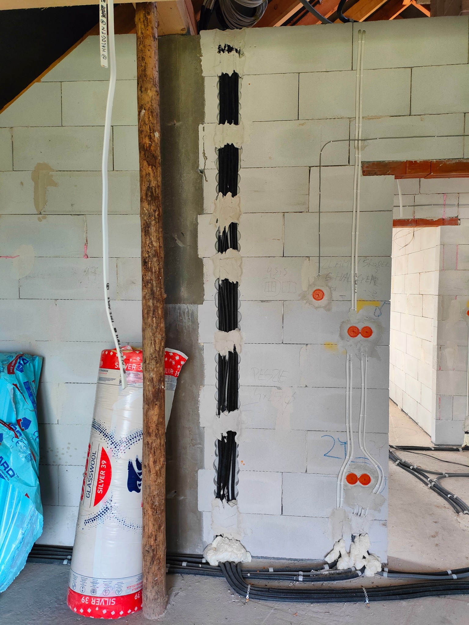 Ściana z betonu komórkowego w trakcie prac instalacyjnych: pionowe bruzdy z czarnymi rurkami peszel wypełnione pianką, kable elektryczne prowadzone po ścianie, puszki elektryczne z pomarańczowymi...