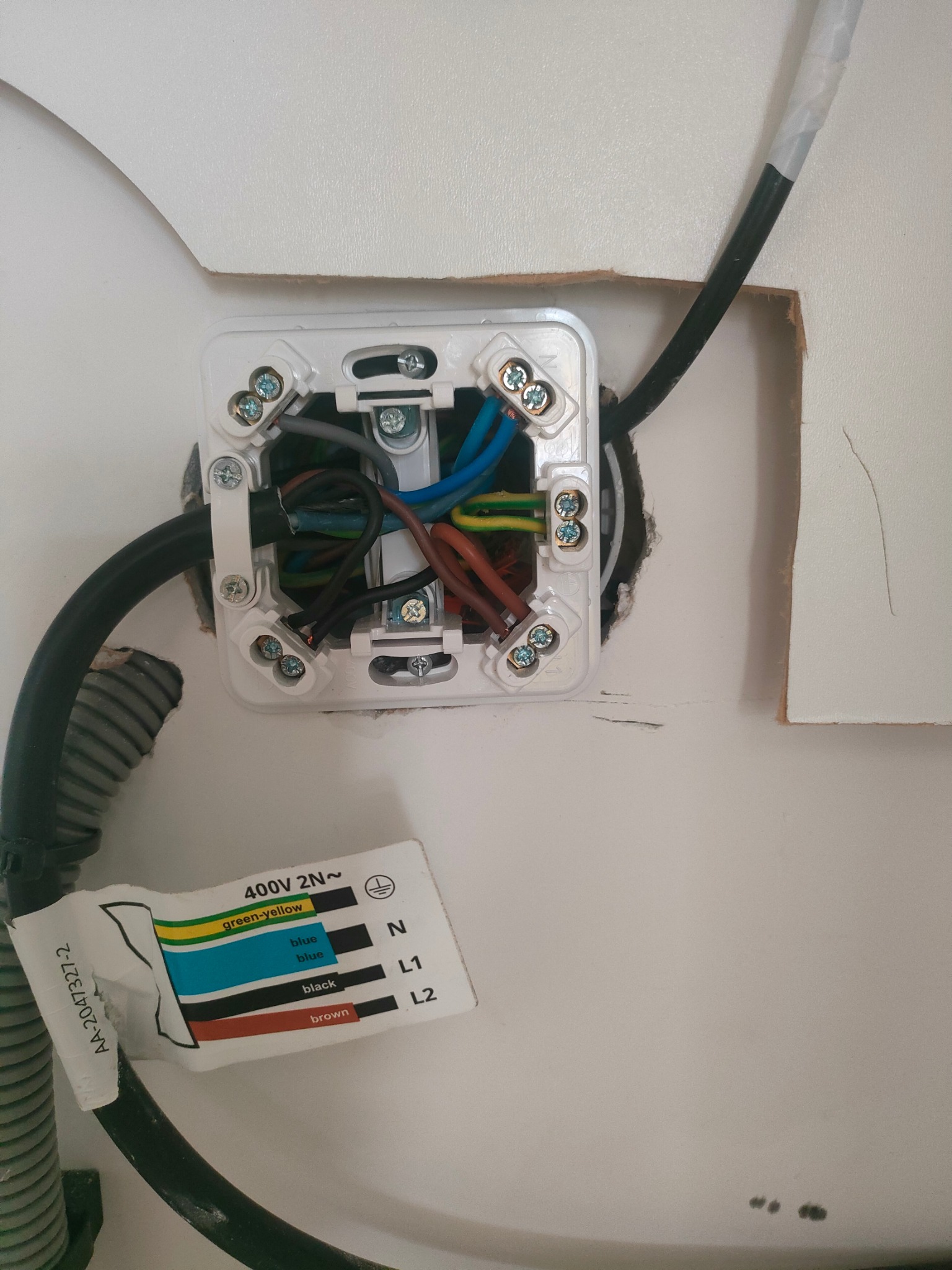 Otwarta puszka elektryczna z podłączonymi przewodami o różnych kolorach, widoczna etykieta z oznaczeniami przewodów, instalacja w ścianie z widocznymi uszkodzeniami.